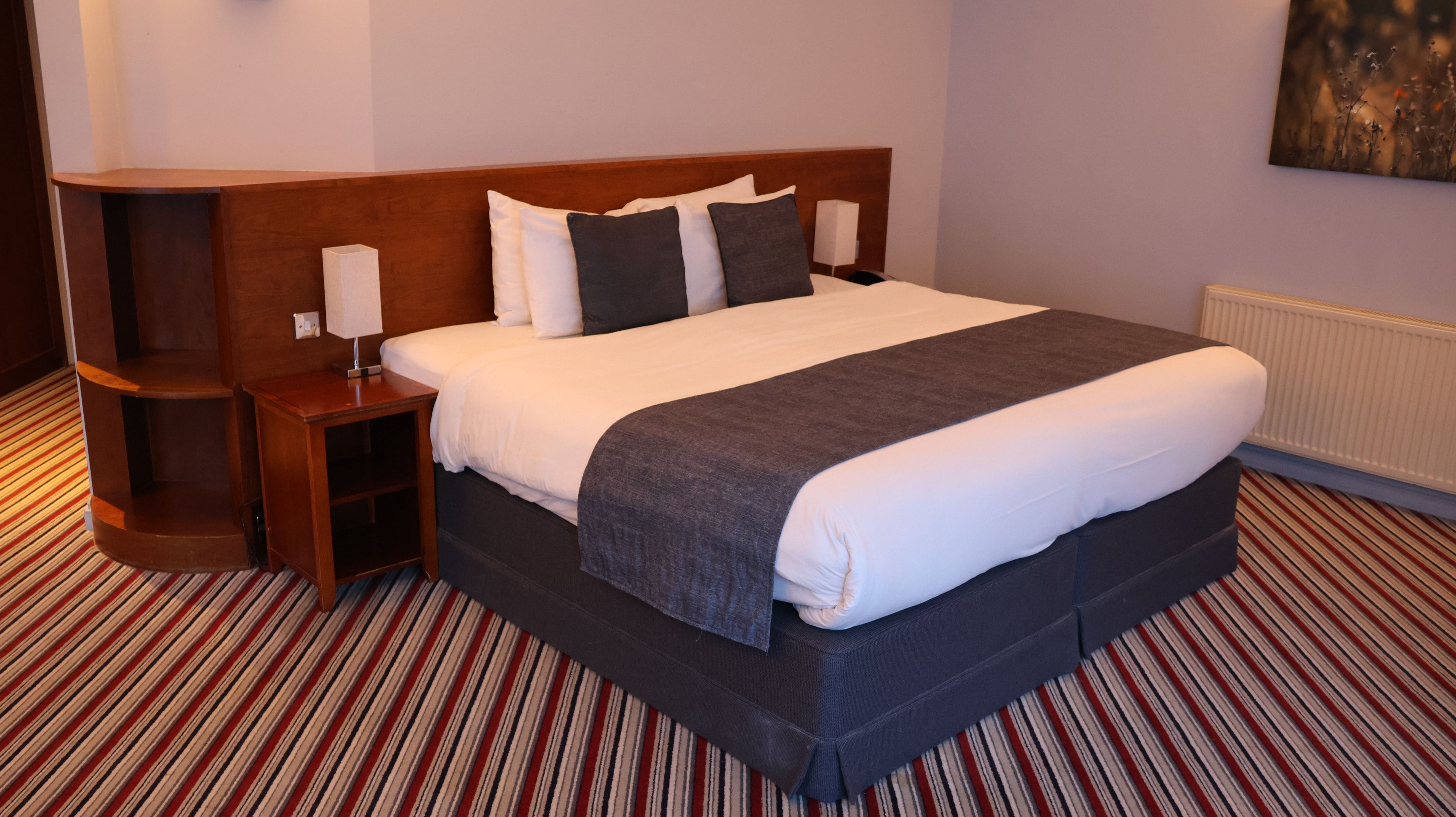 A hotel room with a large bed, a wooden headboard, a bedside table with a lamp, and a striped carpet.
