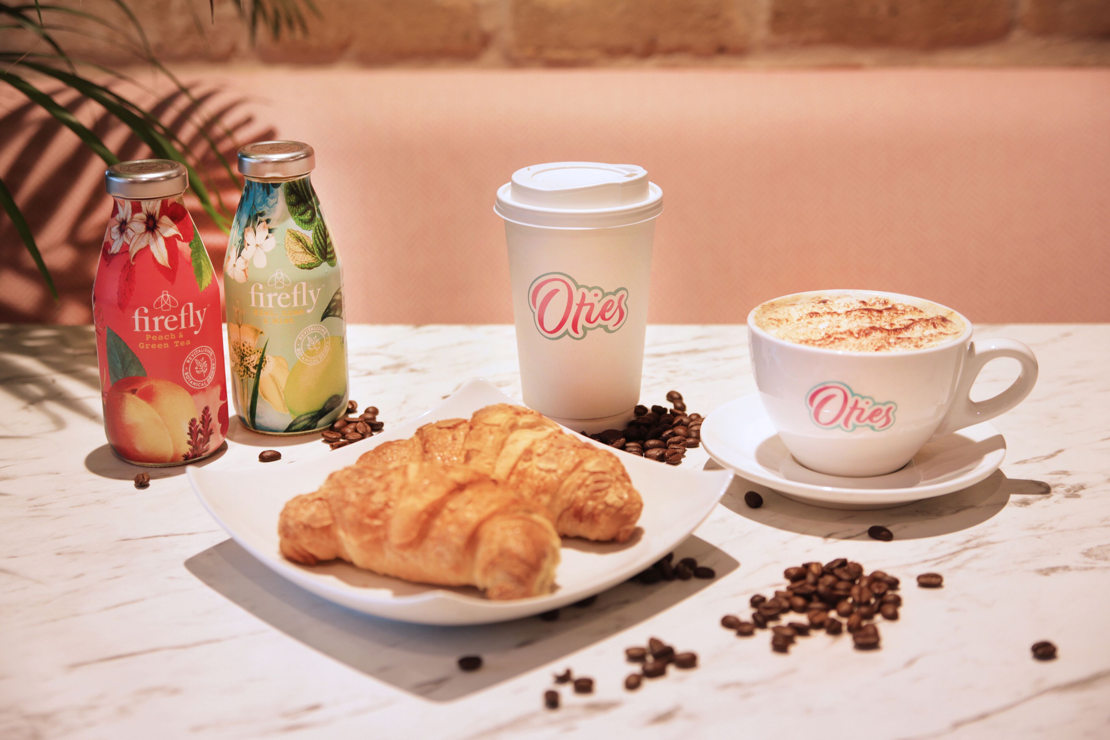 Taula amb un croissant, dues ampolles de te fred, una tassa de cafè i grans de cafè dispersos.