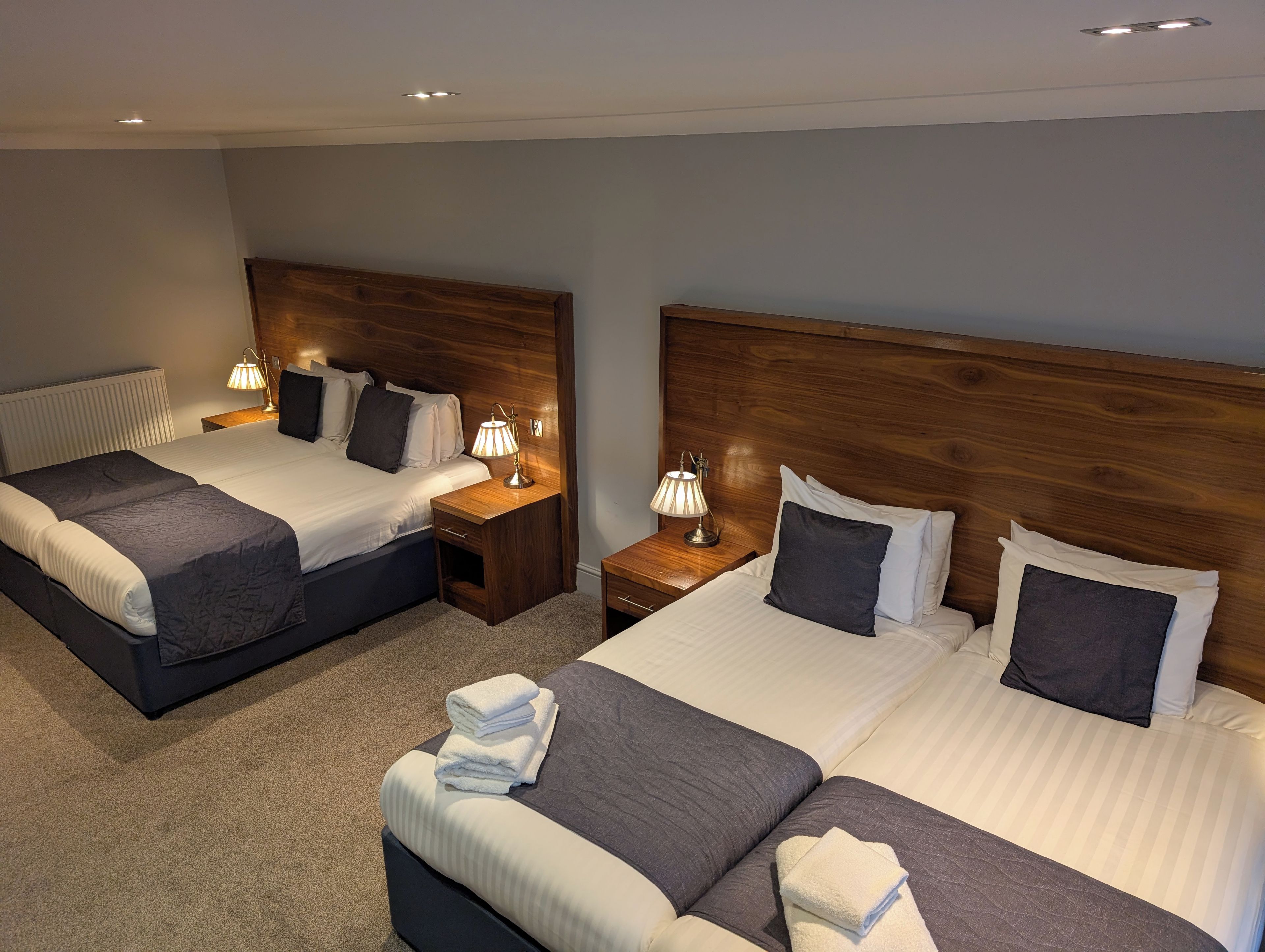A hotel room with two beds, each with a wooden headboard, bedside tables with lamps, and a radiator.