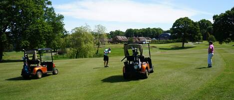 Un campo de golf con dos carritos de golf y una persona con camisa rosa sobre el green.
