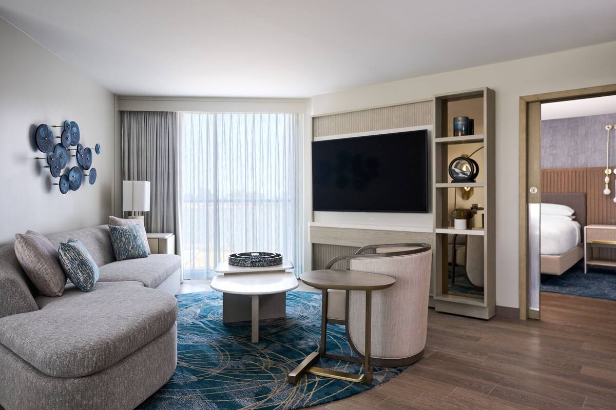 Una habitación de hotel moderna con un sofá gris, una mesa redonda de centro, un televisor de pantalla plana y una estantería.