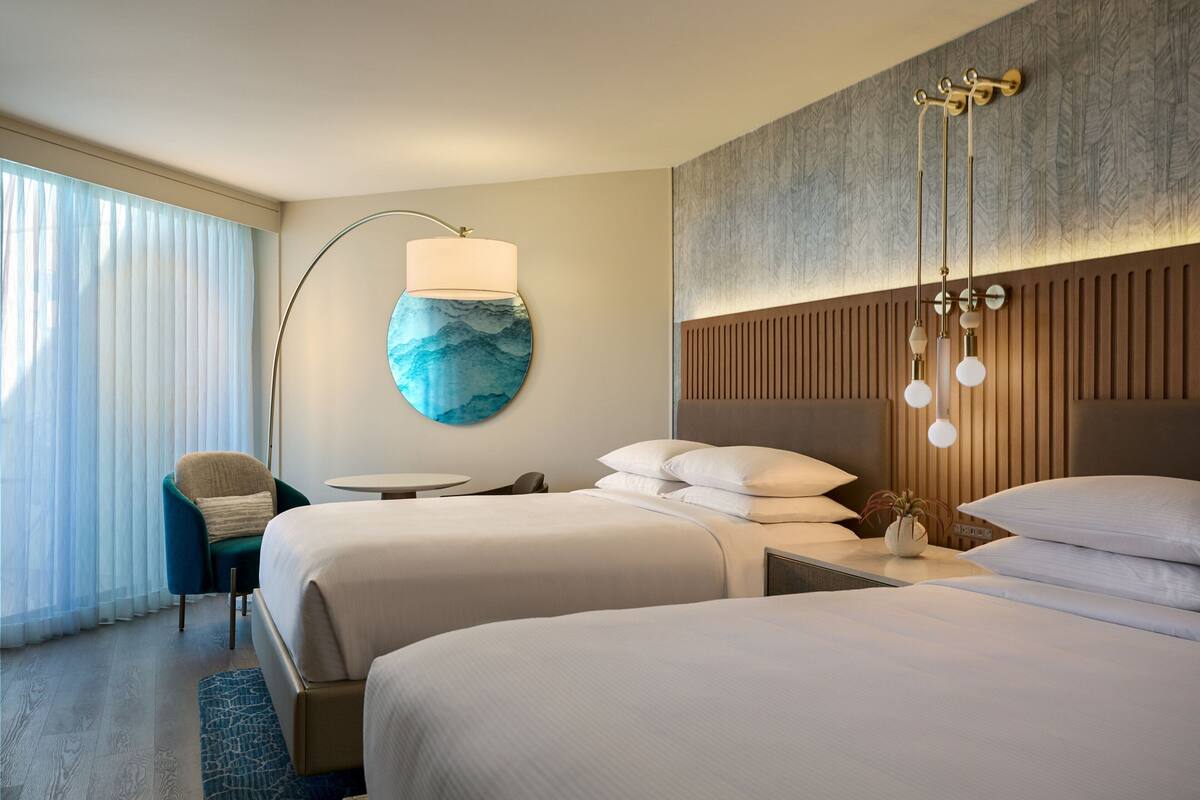 Habitación de hotel con dos camas, una lámpara de pared redonda, una mesita, una silla y un ventanal con cortinas.