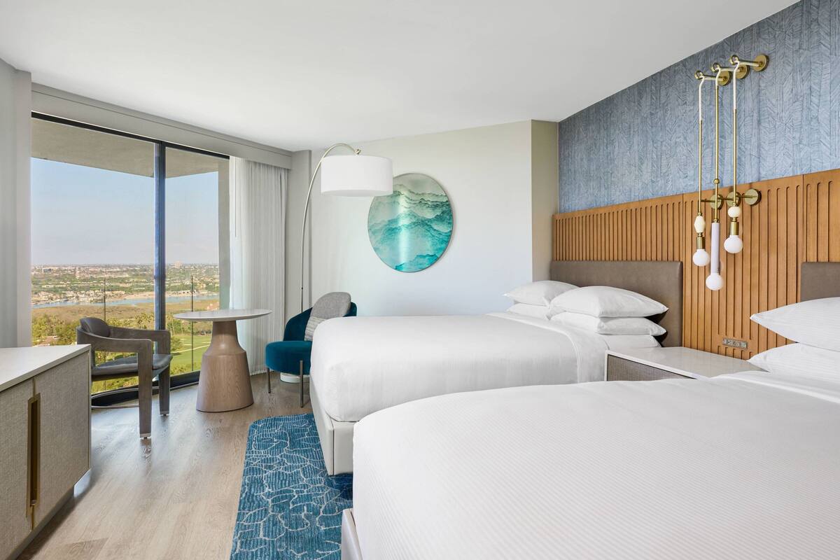 Habitación de hotel con dos camas, un escritorio, una silla, una mesa redonda y un ventanal con vistas a la ciudad.