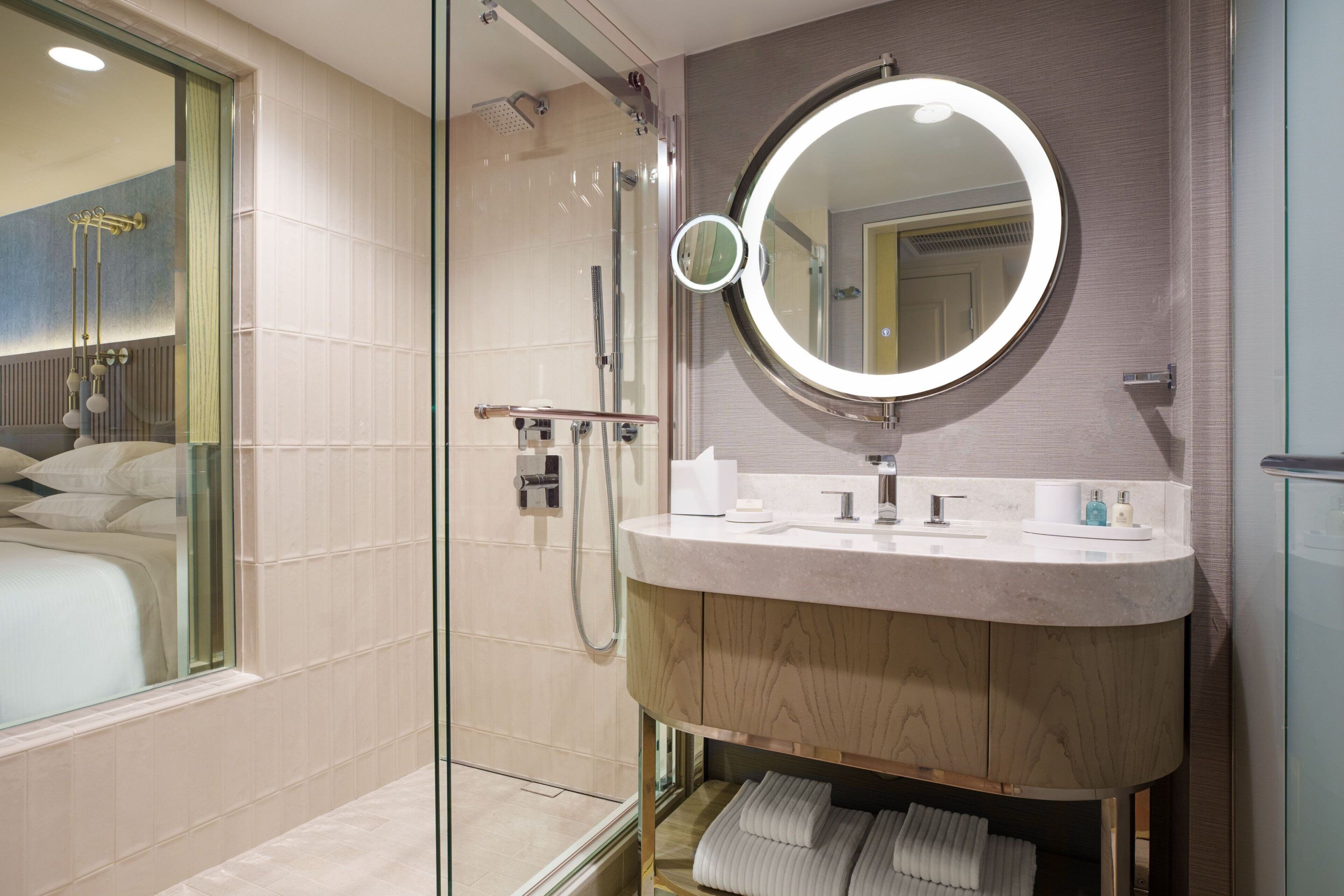 A modern bathroom with a glass-enclosed shower, a round mirror, and a wooden vanity with a marble countertop.