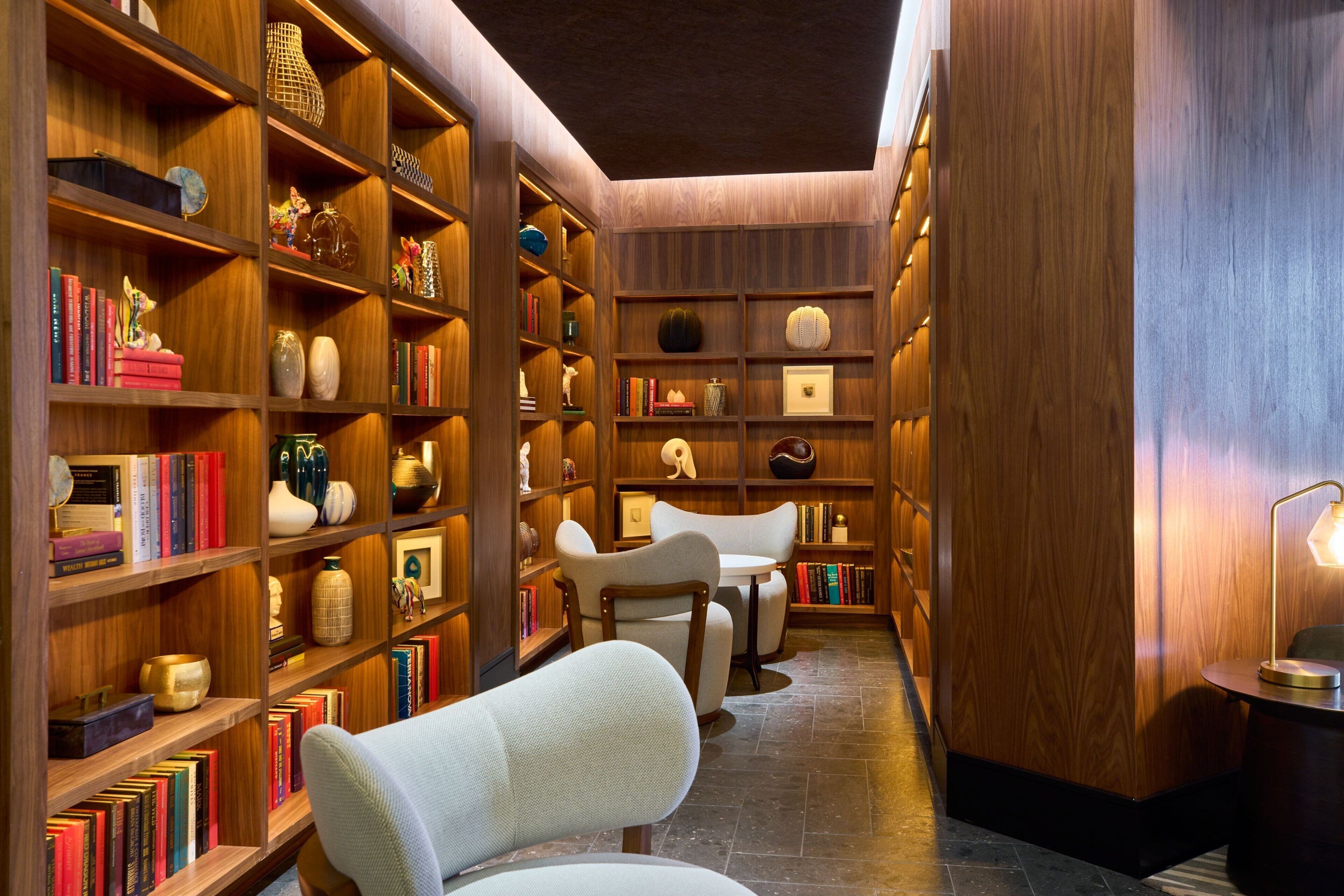 Um corredor estreito e acolhedor, iluminado com luz quente, com estantes de madeira repletas de livros e objetos decorativos, que leva a um espaço de estar com duas cadeiras e uma mesa.