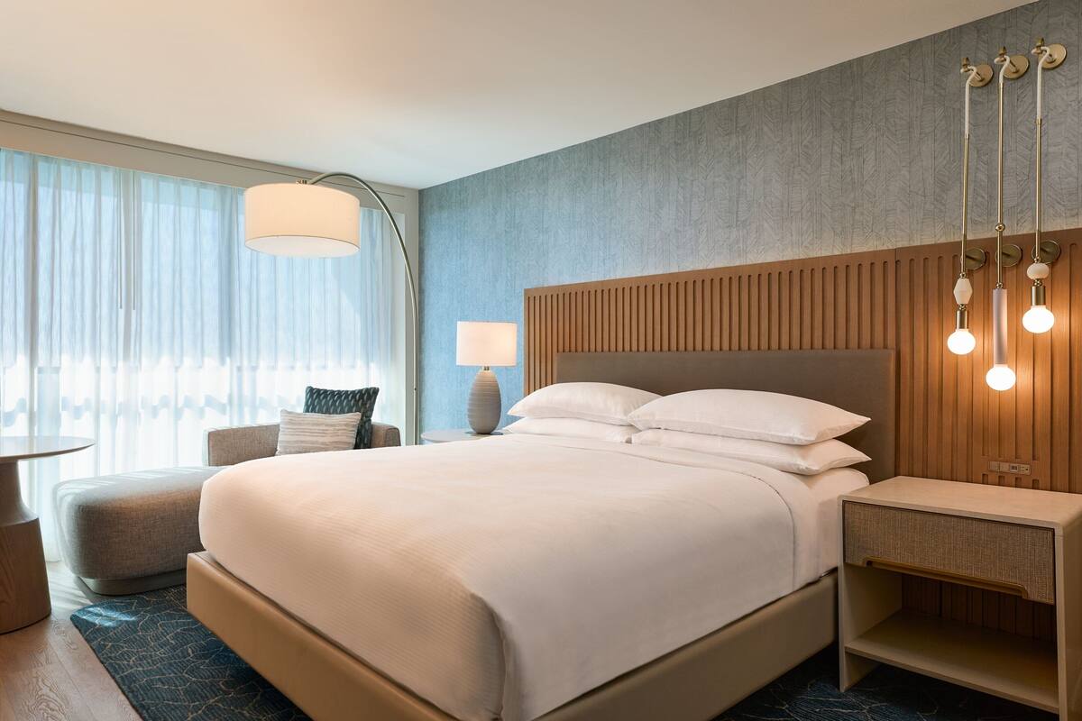 Una habitación de hotel moderna con una cama grande, una mesita de noche con lámpara, un pequeño rincón de descanso y un ventanal con cortinas traslúcidas.