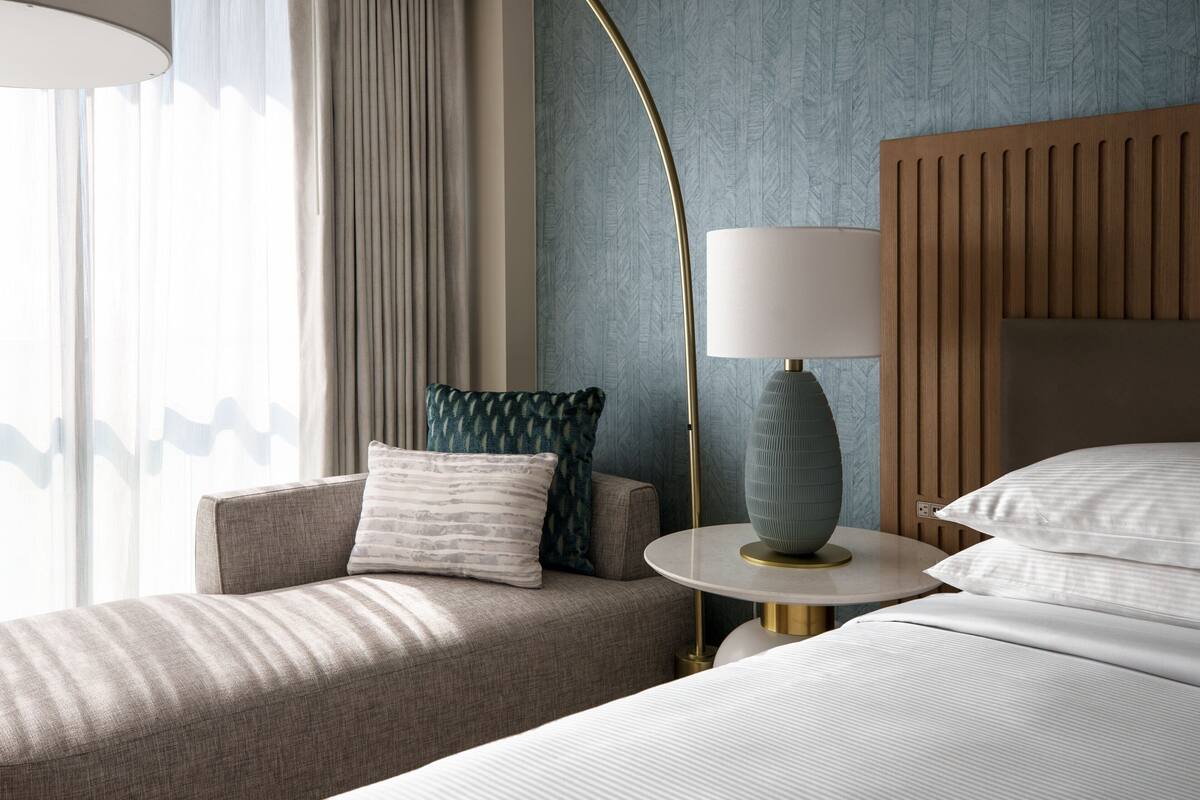 Una habitación de hotel con sofá, una mesa redonda con lámpara, cama con almohadas y cabecero de madera.
