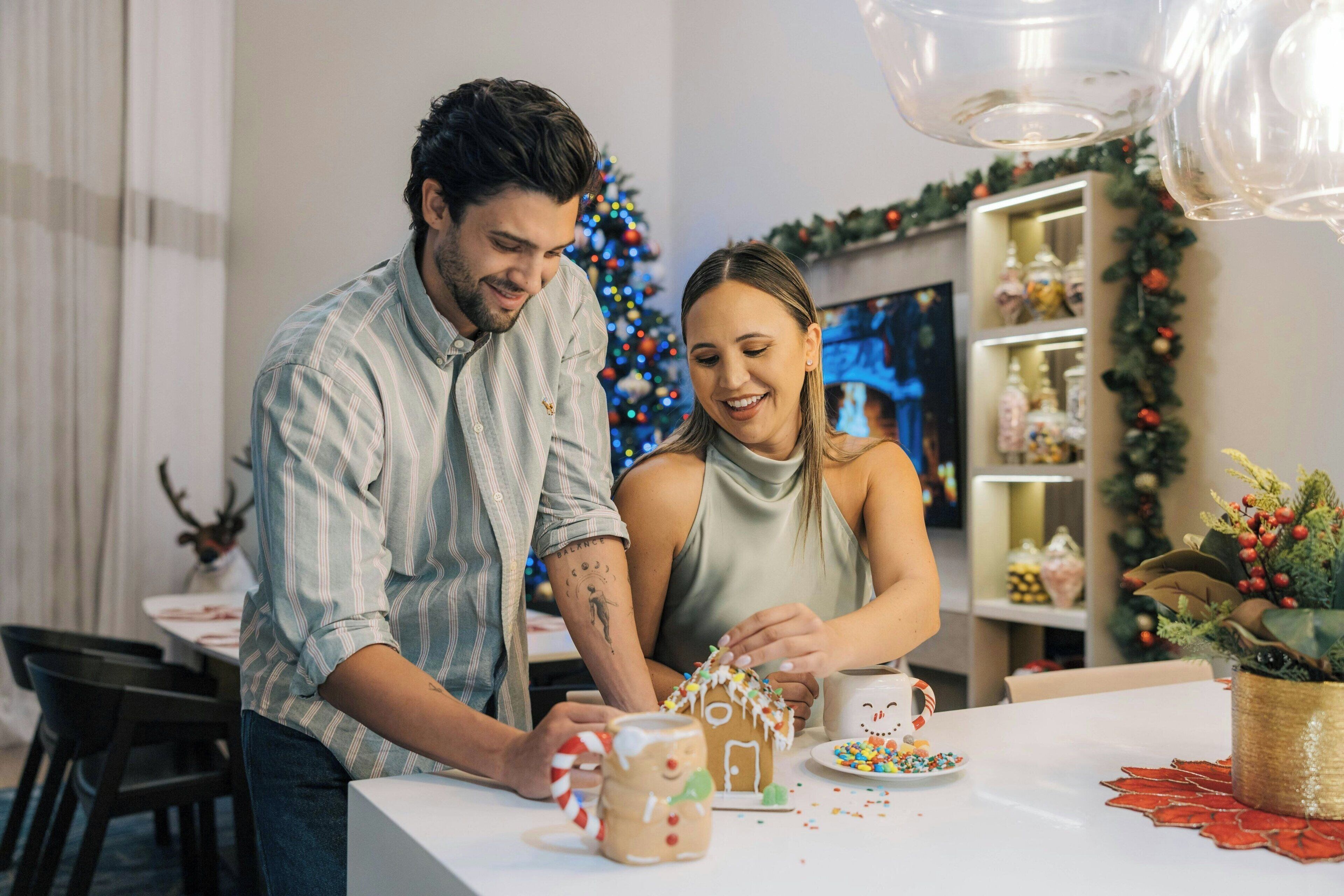A couple decorating a gingerbread house in a festive room with a Christmas tree and holiday decorations.
