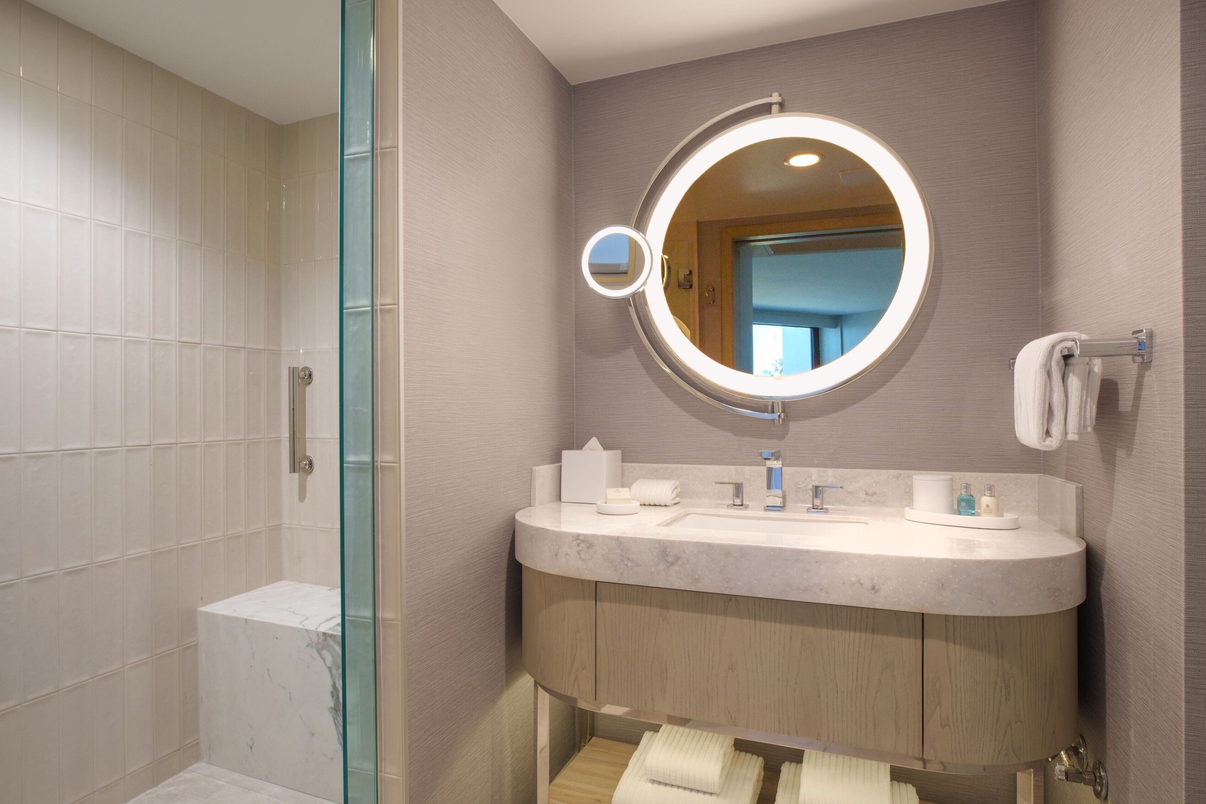 Un baño moderno con cabina de ducha de vidrio, lavamanos de mármol y espejo redondo con iluminación integrada.
