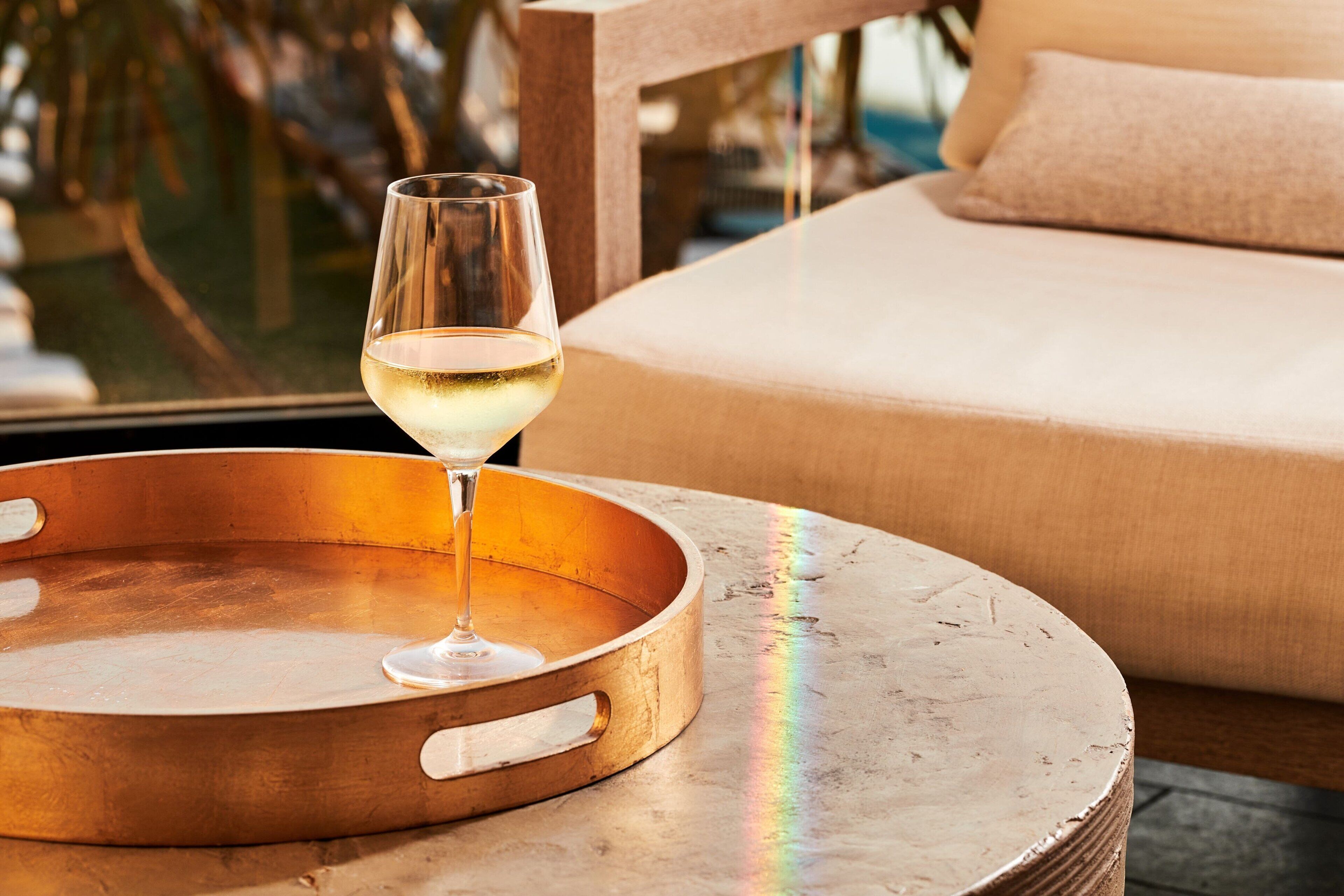 A glass of white wine on a copper tray, a beige sofa, and a view of the outdoors.