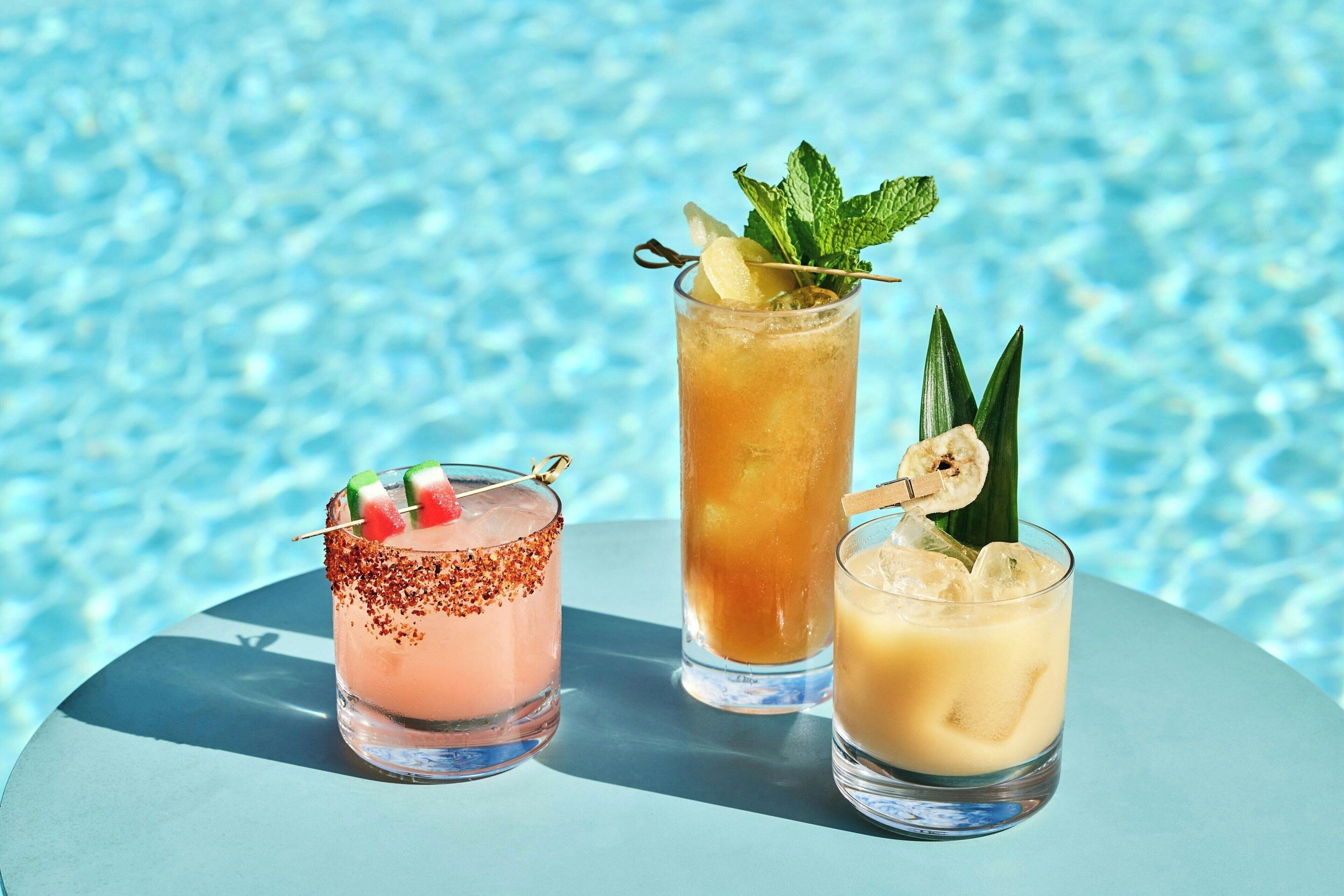 Three cocktails with garnishes, placed on a table by a pool.