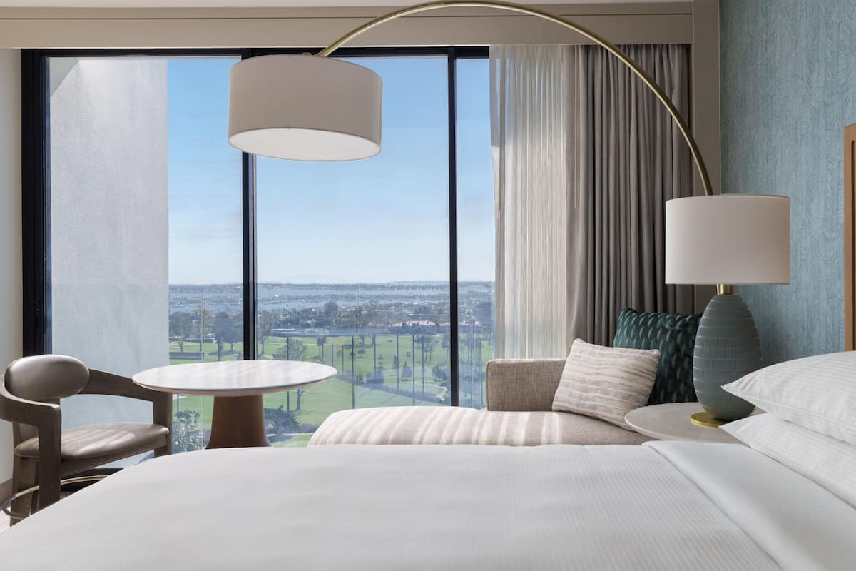 Habitación de hotel con una cama grande, una mesa redonda, una silla y vistas a un paisaje urbano a través de amplios ventanales.