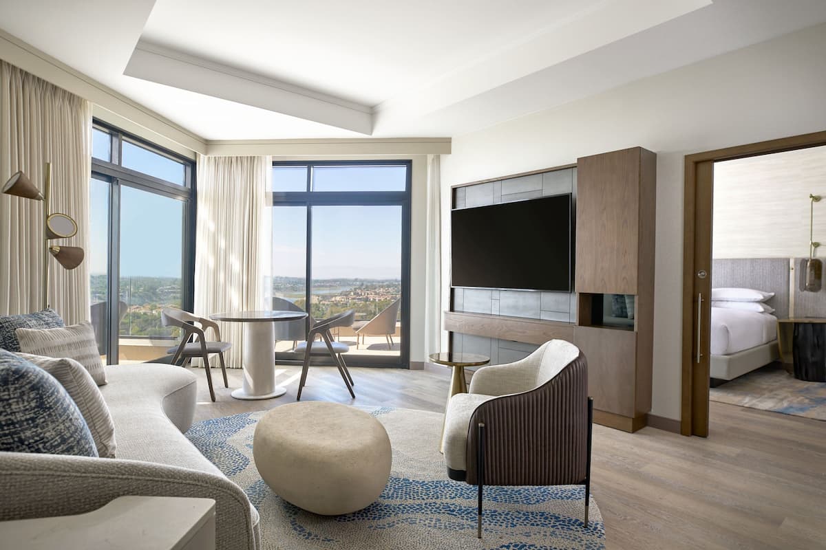 Una habitación de hotel moderna con una cama grande, televisión, balcón con vistas y una zona de estar con mesa y sillas.