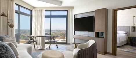 Ein modernes Hotelzimmer mit einem großen Bett, einem Fernseher, einem Balkon mit Aussicht und einer Sitzecke mit Tisch und Stühlen.