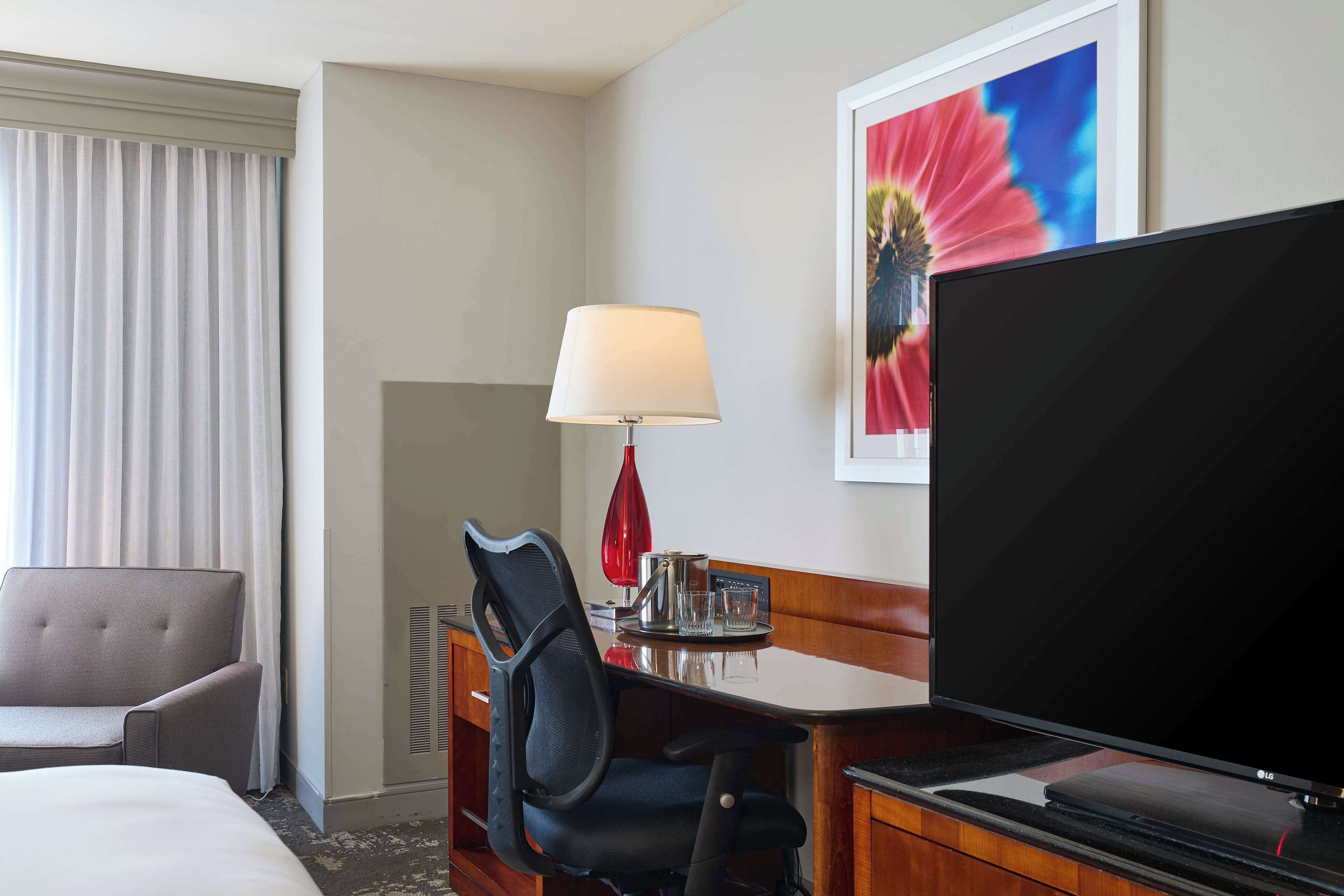 Une chambre d’hôtel avec un lit, un bureau avec une télévision, une chaise, une lampe et une fenêtre avec des rideaux.