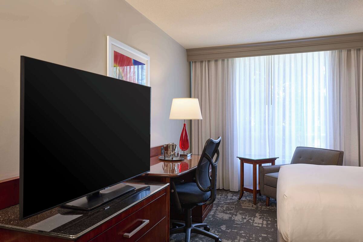 Une chambre d’hôtel équipée d’un téléviseur à écran plat, d’un bureau avec une lampe, d’une chaise, d’un lit et d’une fenêtre avec des rideaux.