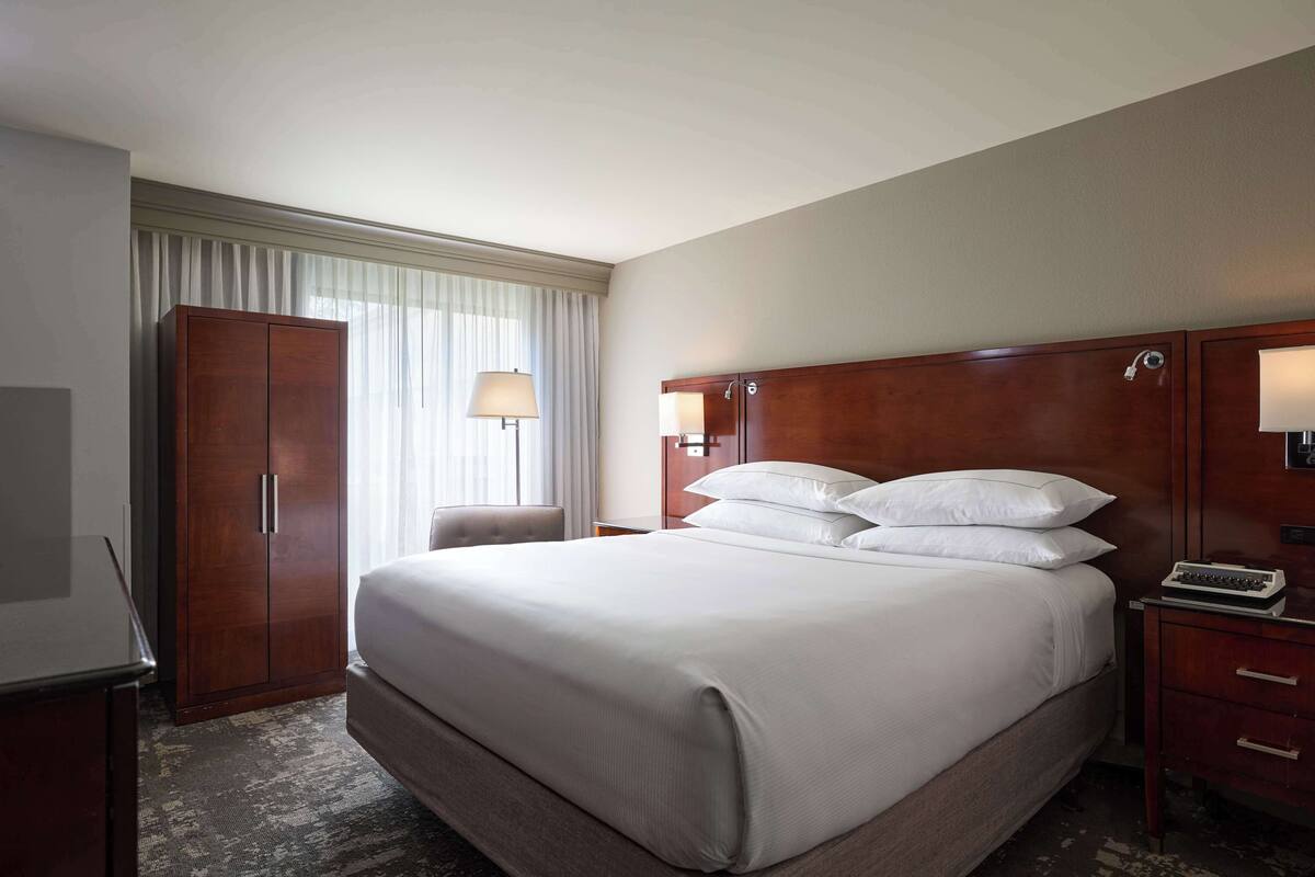 Une chambre d’hôtel avec un grand lit, une armoire, un bureau avec une lampe et une fenêtre avec des rideaux.