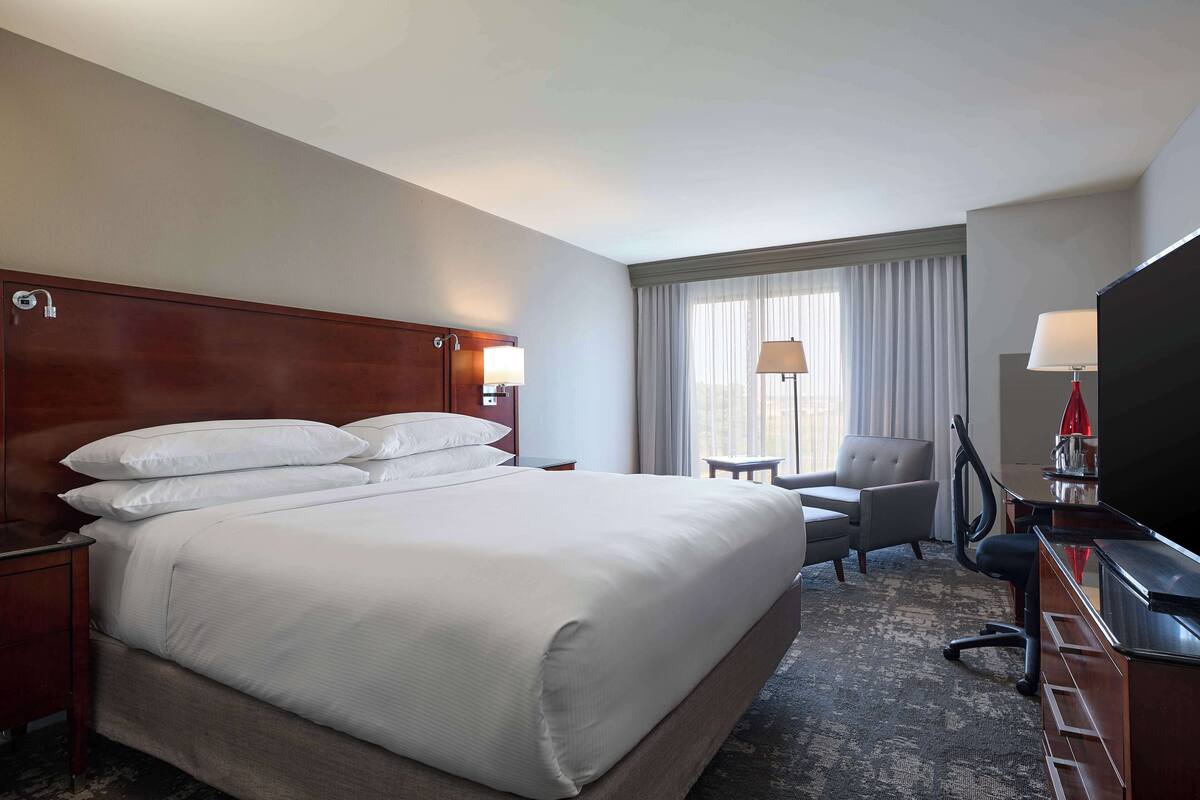 Une chambre d’hôtel avec un grand lit, un bureau avec une télévision, une chaise et une fenêtre avec des rideaux.