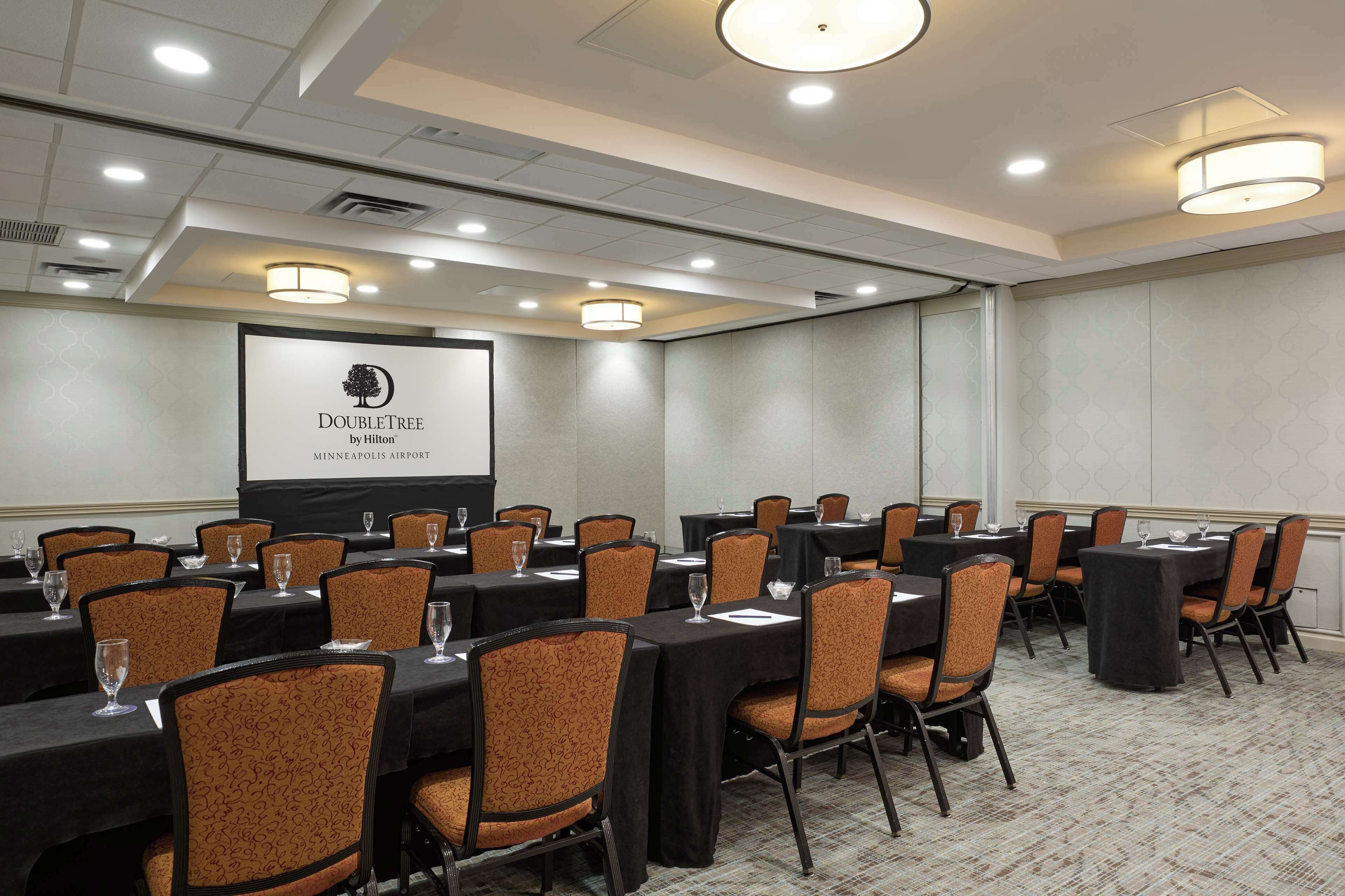 A conference room with multiple tables and chairs, a projector screen displaying the DoubleTree by Hilton logo, and a carpeted floor.