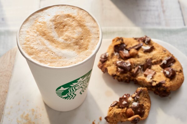 A Starbucks cup with a frothy top and a cookie with chocolate chips on a white plate.