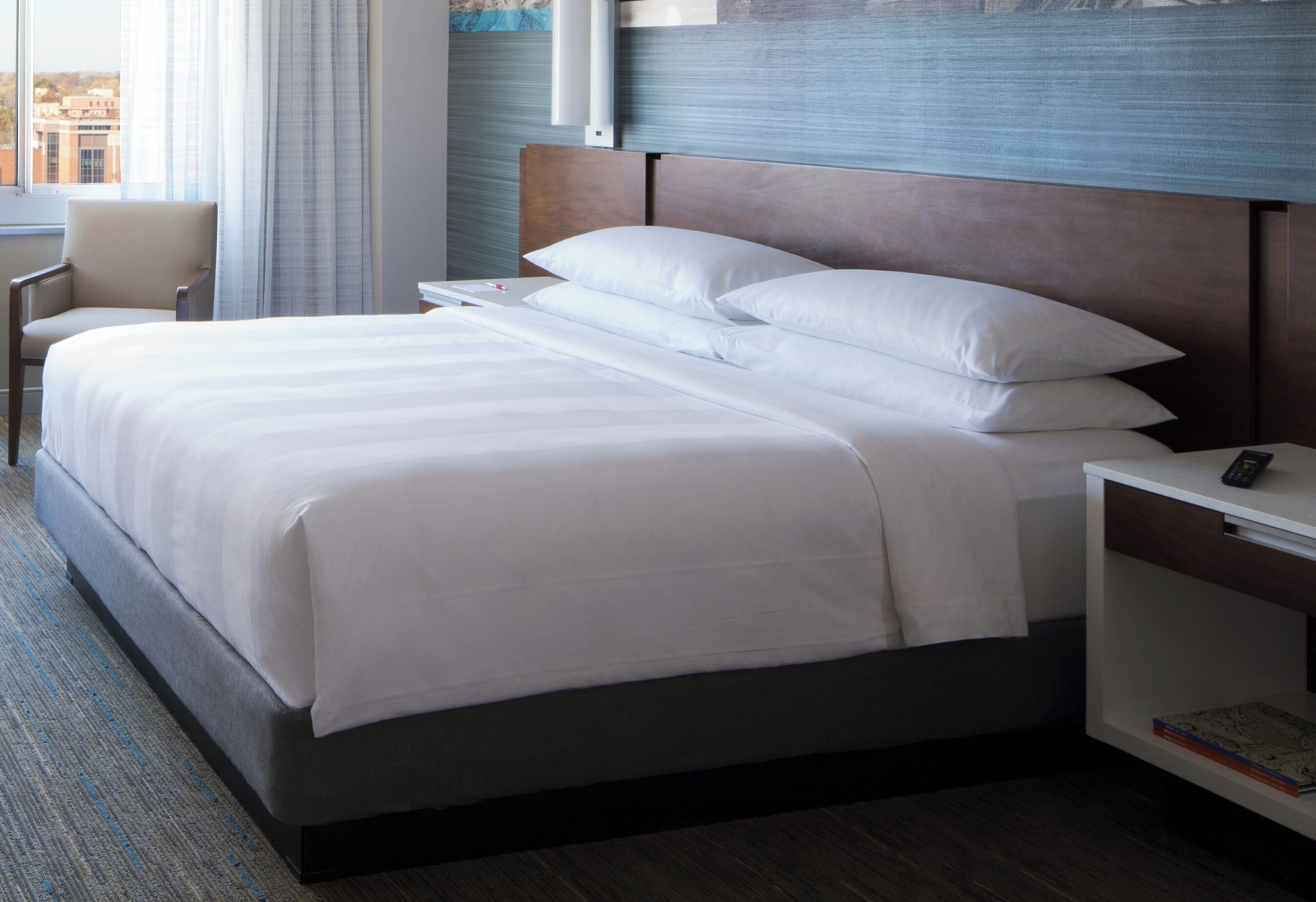 Ein Hotelzimmer mit einem Bett, einem Sessel, einem Nachttisch mit Fernbedienung und einem Fenster mit Vorhängen.