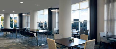 Un comedor moderno de hotel con varias mesas y sillas, grandes ventanales que ofrecen vistas a la ciudad y piso alfombrado.