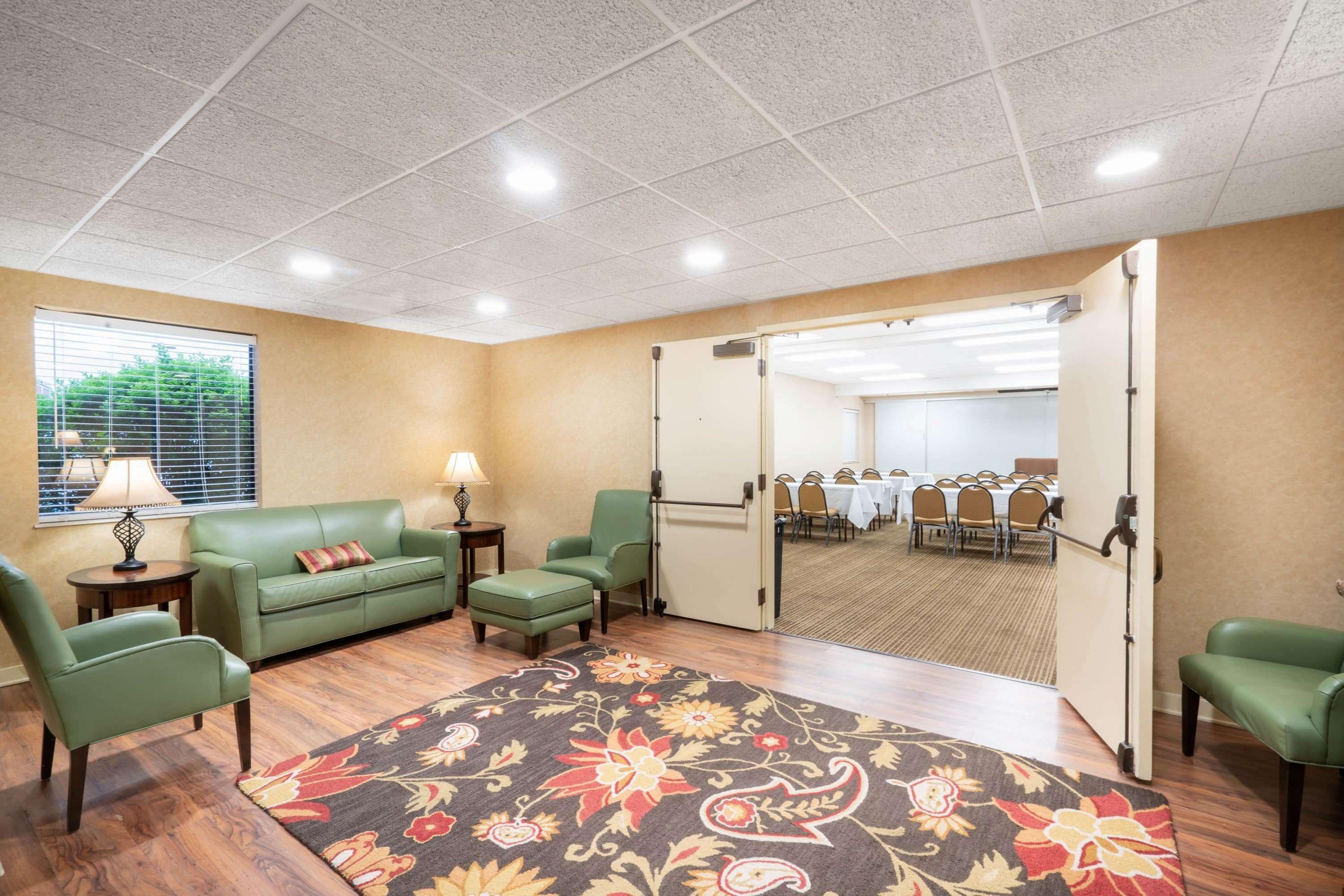 A hotel room with a green sofa, a matching armchair, a small table with a lamp, a patterned rug, and a view of a conference room through a sliding door.
