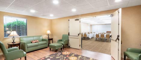 A hotel room with a green sofa, a matching armchair, a small table with a lamp, a patterned rug, and a view of a conference room through a sliding door.