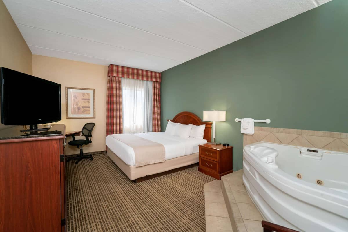 A hotel room with a bed, a desk, a television, a chair, a desk lamp, a bedside table, and a bathtub.