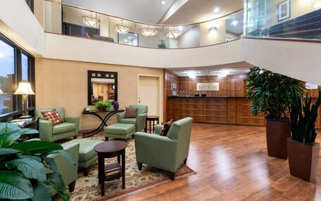 A spacious lobby with a high ceiling, a central seating area with green upholstered chairs, a wooden reception desk, and a large potted plant.