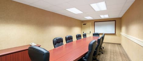 A conference room with a long wooden table, six black chairs, a whiteboard, and a projector screen.