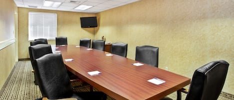 Une salle de conférence avec une longue table en bois, des fauteuils en cuir noir, un téléviseur à écran plat fixé au mur et un sol recouvert de moquette.