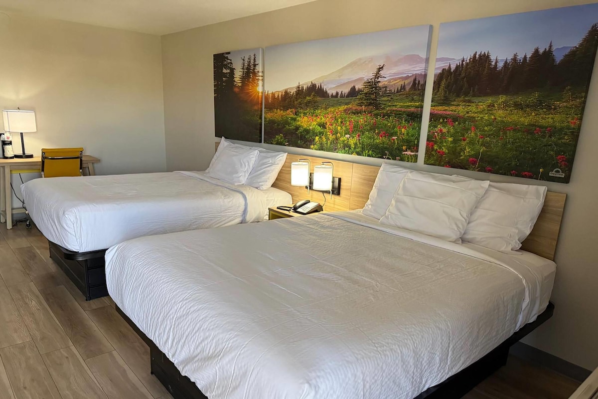 A hotel room with two beds, a desk with a lamp, and a large landscape painting on the wall.