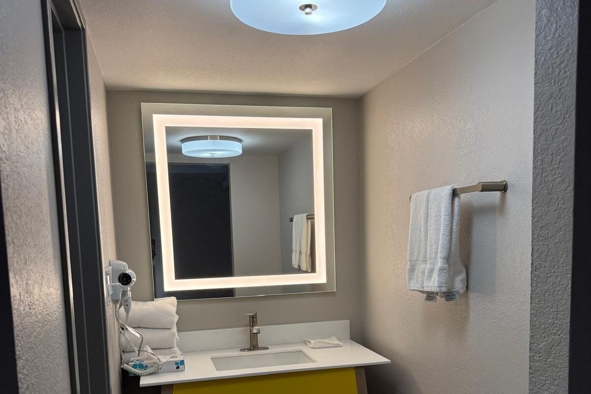 A modern bathroom with a white sink, yellow vanity, and a mirror with a light above it.