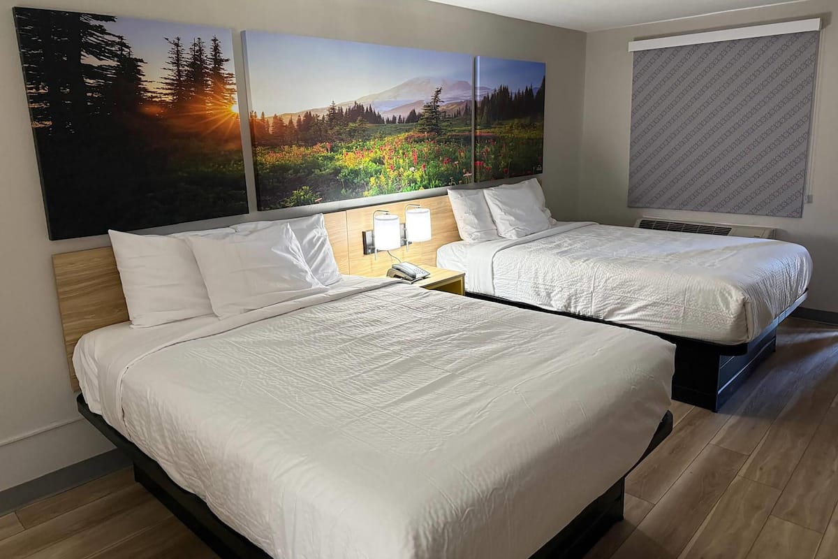 A hotel room with two beds, a wooden headboard, a nightstand with a lamp, and a large window with blinds.