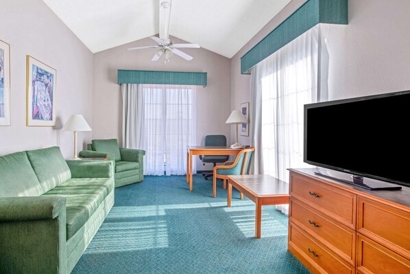 A hotel room with a green sofa, a wooden desk, a television, and a ceiling fan.