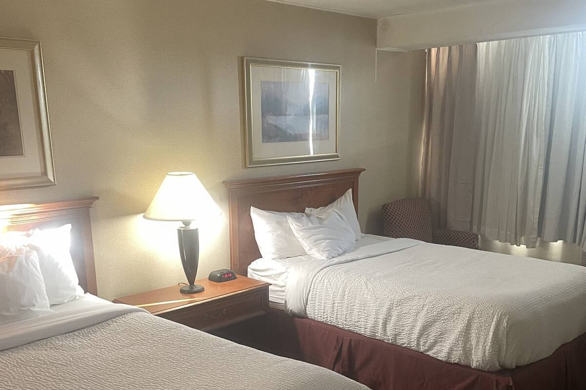 A hotel room with two beds, a nightstand with a lamp, a framed picture on the wall, and a window with curtains.