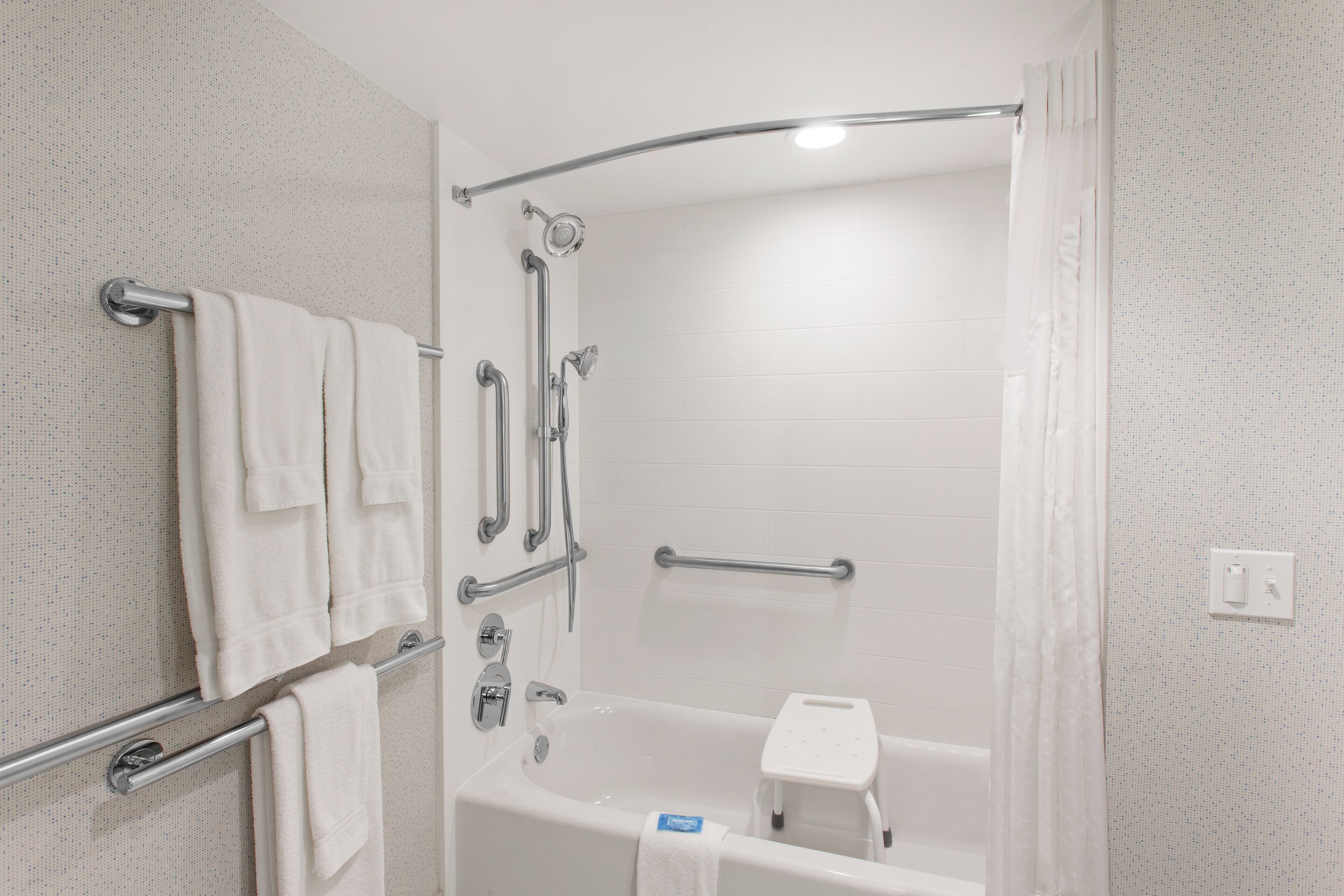 Un bagno moderno con doccia a ingresso libero, maniglie di sostegno, una vasca da bagno bianca e uno sgabello.