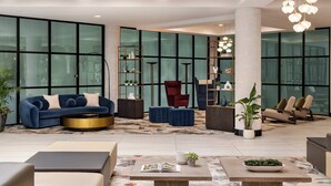 A spacious lobby with a blue sofa, red armchair, and beige chairs. There are potted plants, a coffee table, and a bookshelf.