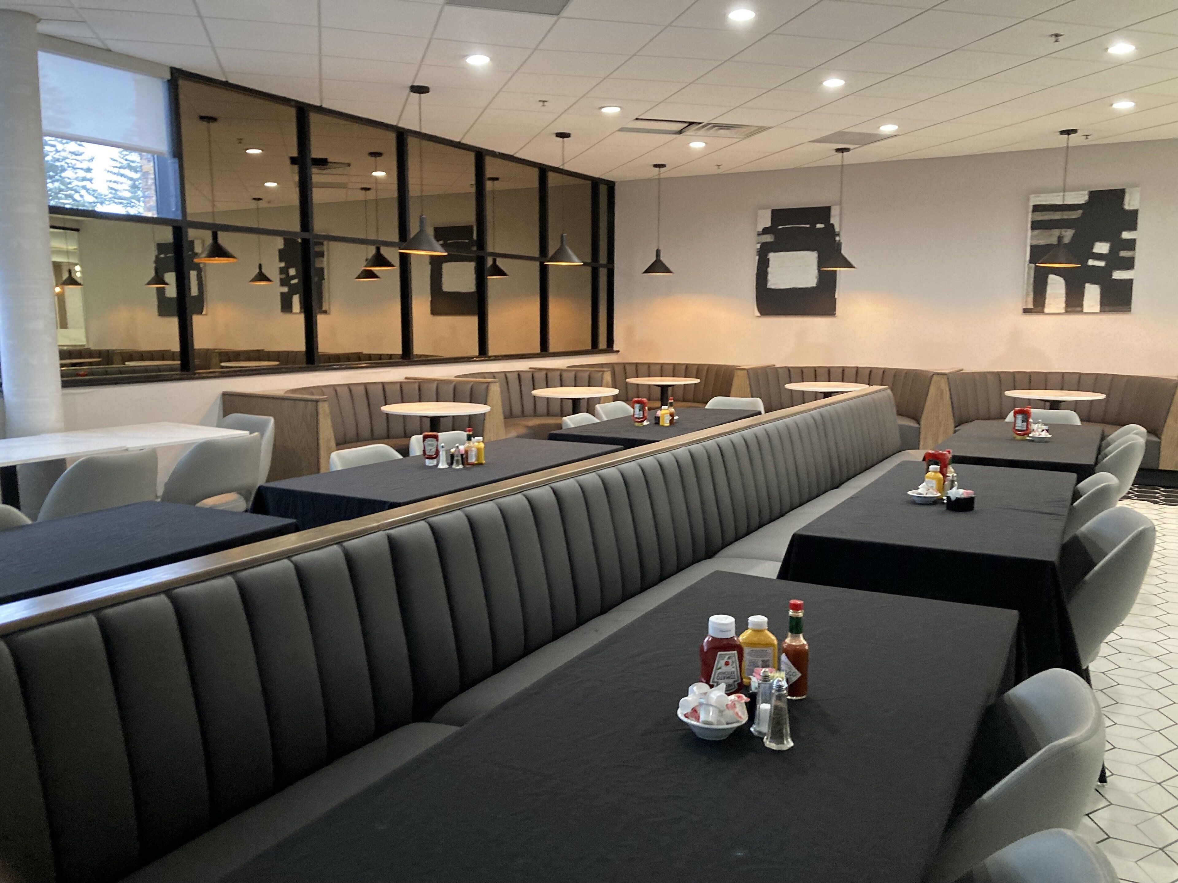 A modern restaurant with booth seating and round tables, featuring a clean, minimalist design and a large window offering a view.