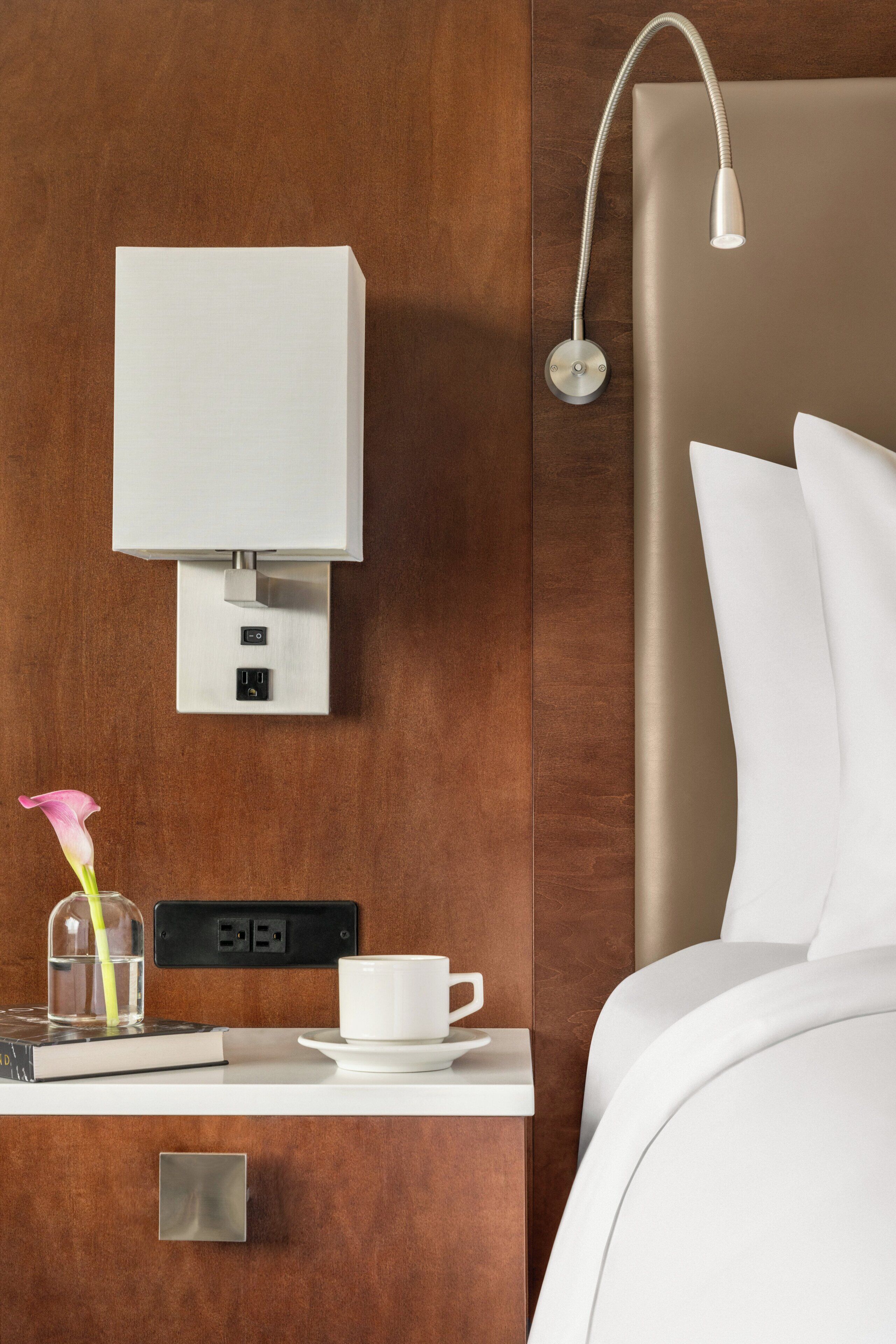 A hotel room with a wooden wall, a bedside table with a lamp, a cup, a book, and a flower vase.