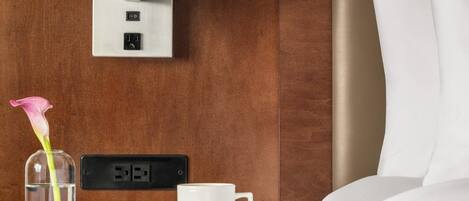 A hotel room with a wooden wall, a bedside table with a lamp, a cup, a book, and a flower vase.