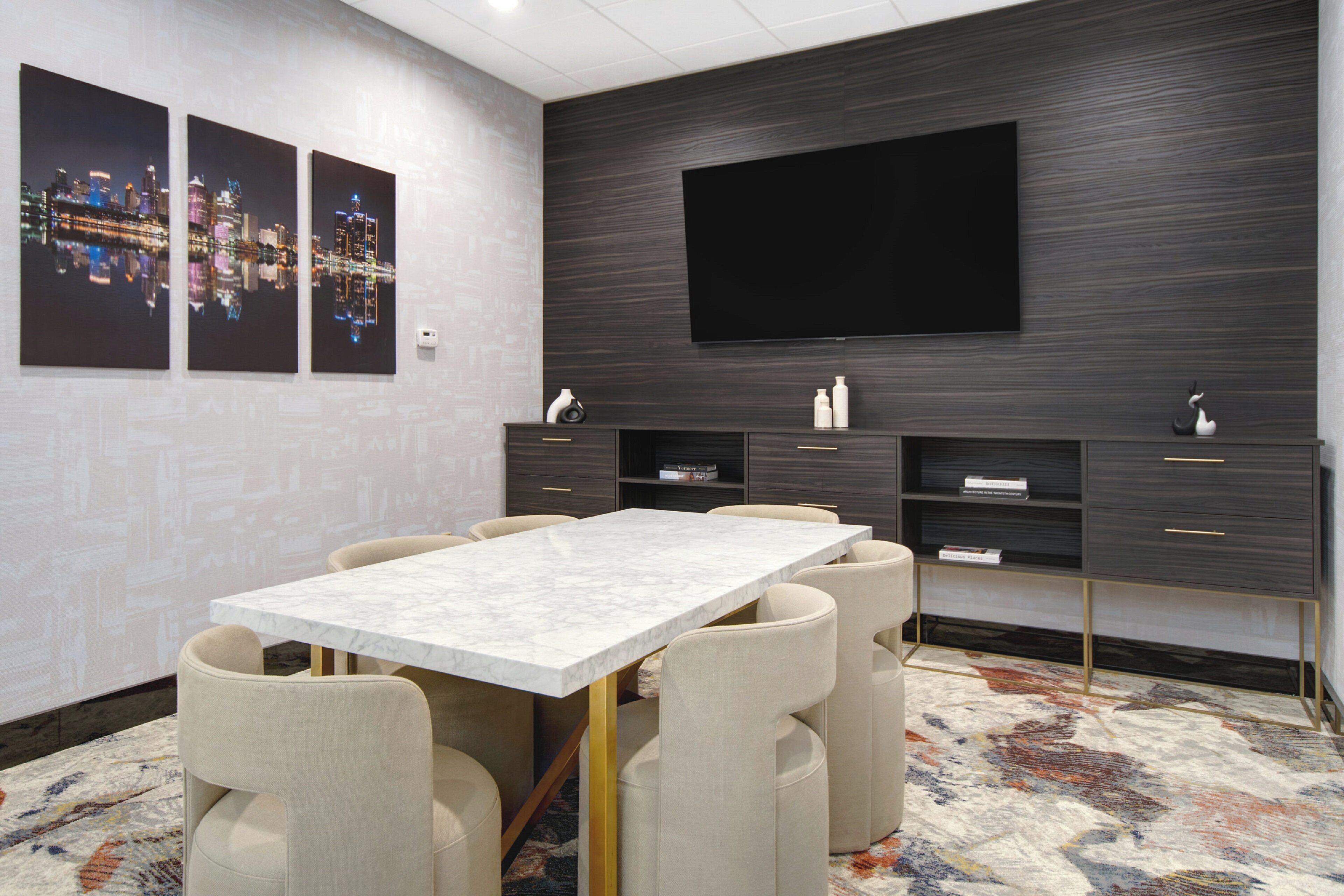 A modern conference room with a marble table, beige chairs, a flat-screen TV, dark wood paneling, and artwork on the walls.