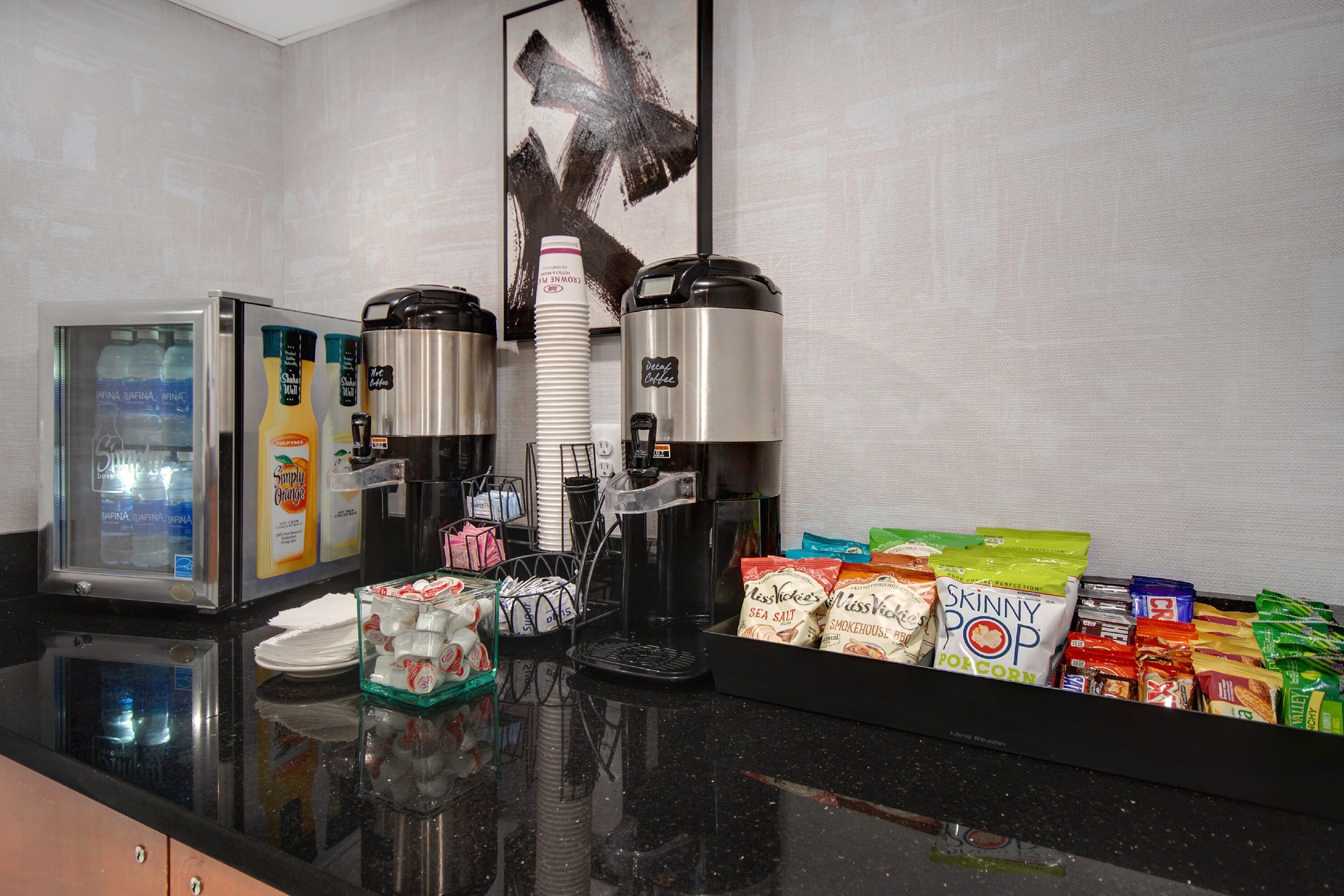 A hotel breakfast bar with a coffee maker, a refrigerator, and various food items including cereal and snacks.
