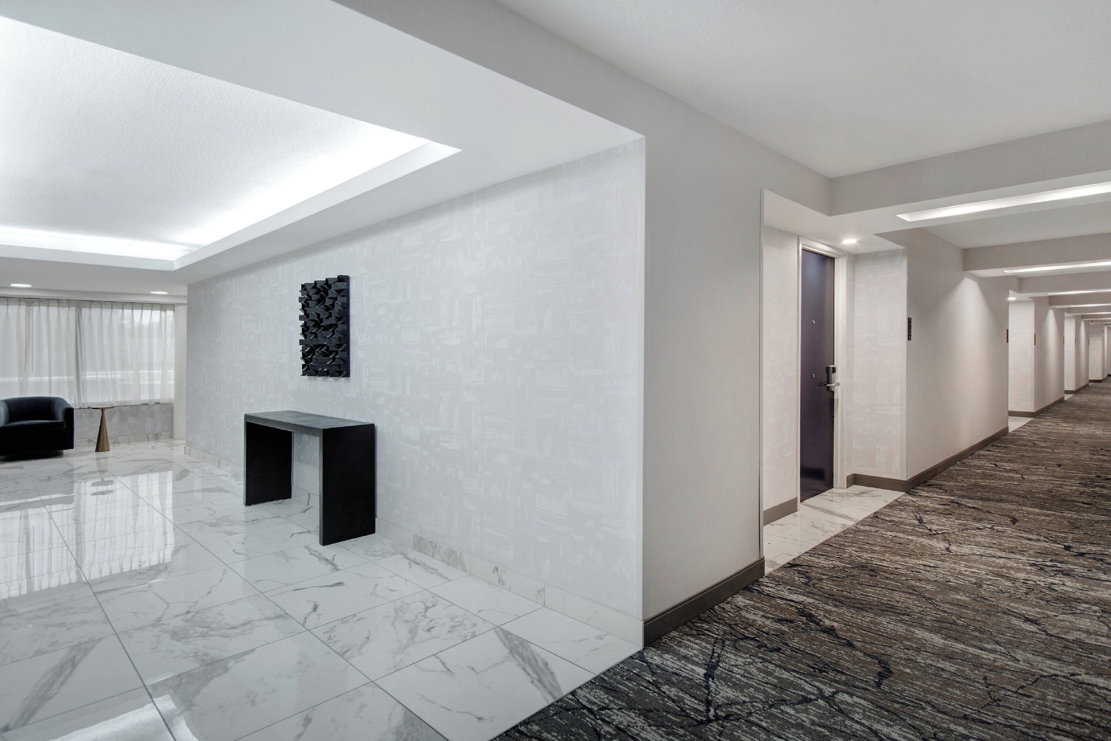A modern hotel lobby with a marble floor, a reception desk, a wall-mounted art piece, and a long corridor with doors.