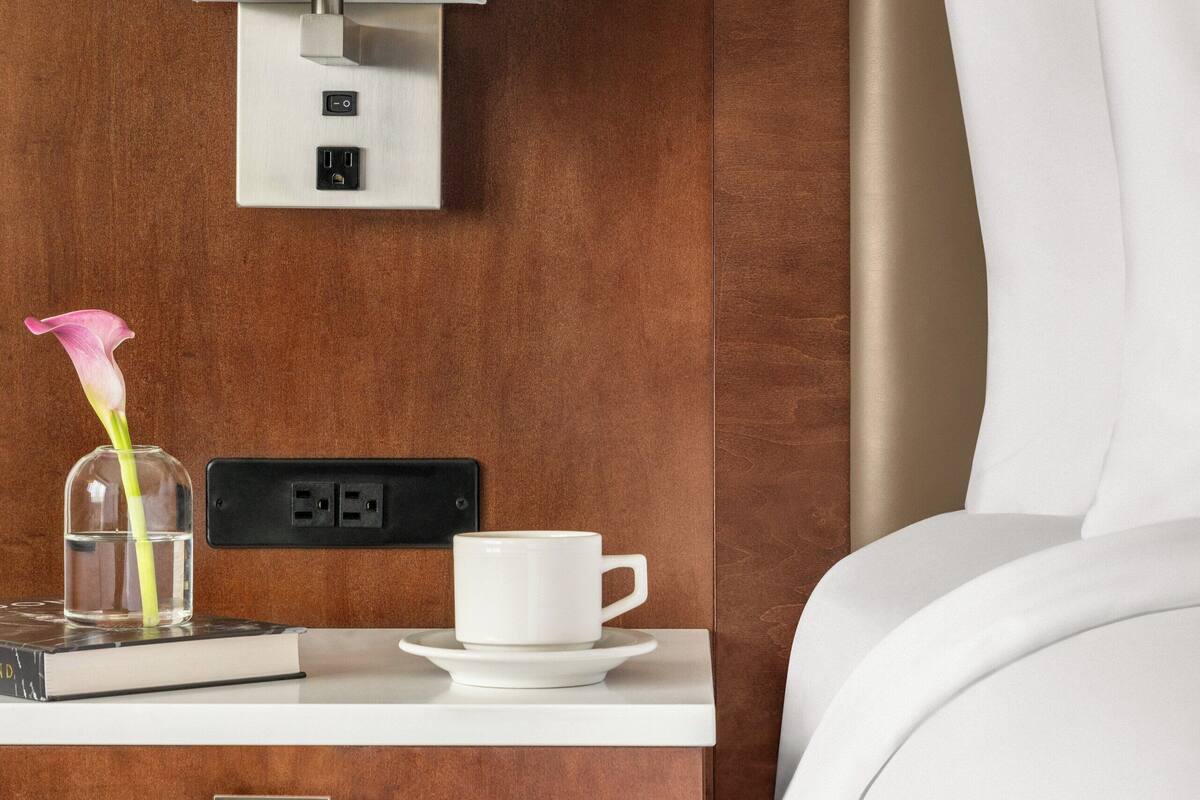 Habitación de hotel con pared de madera, mesita de noche con lámpara, taza, libro y florero.
