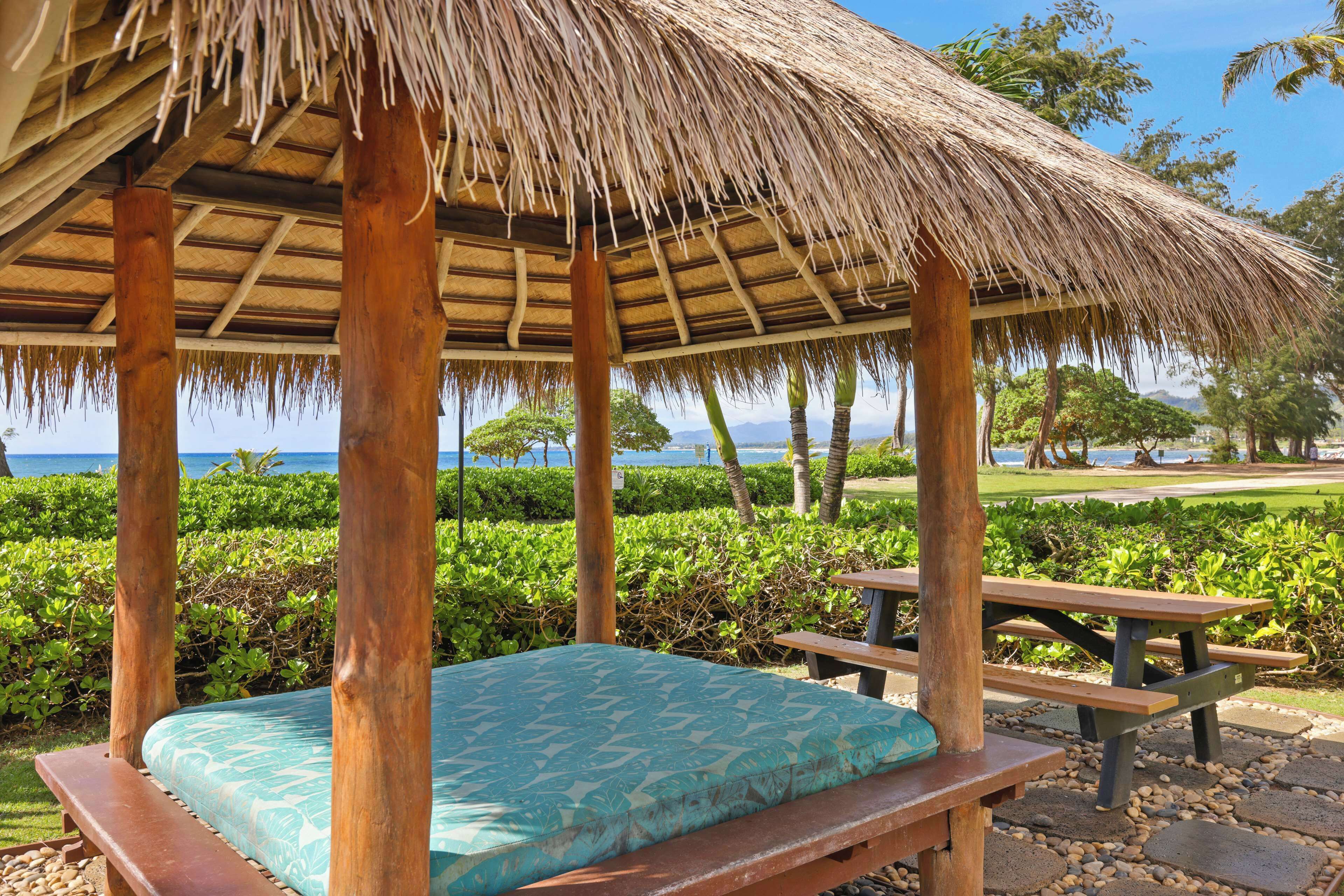 A thatched-roof gazebo with a bed and picnic tables, overlooking a landscaped garden and the ocean.