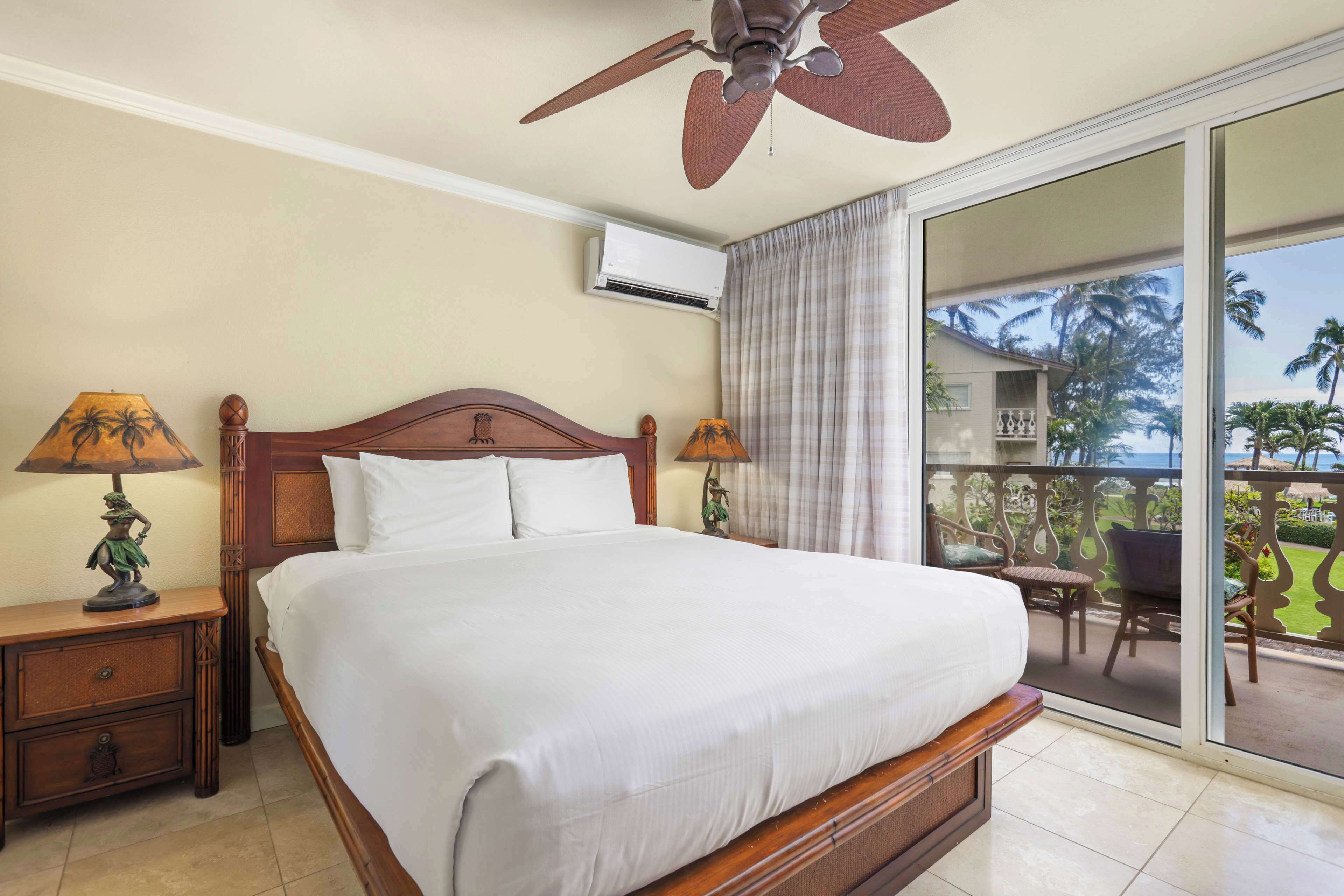 A hotel room with a large bed, two bedside tables with lamps, a ceiling fan, and a sliding glass door leading to a balcony with outdoor furniture.