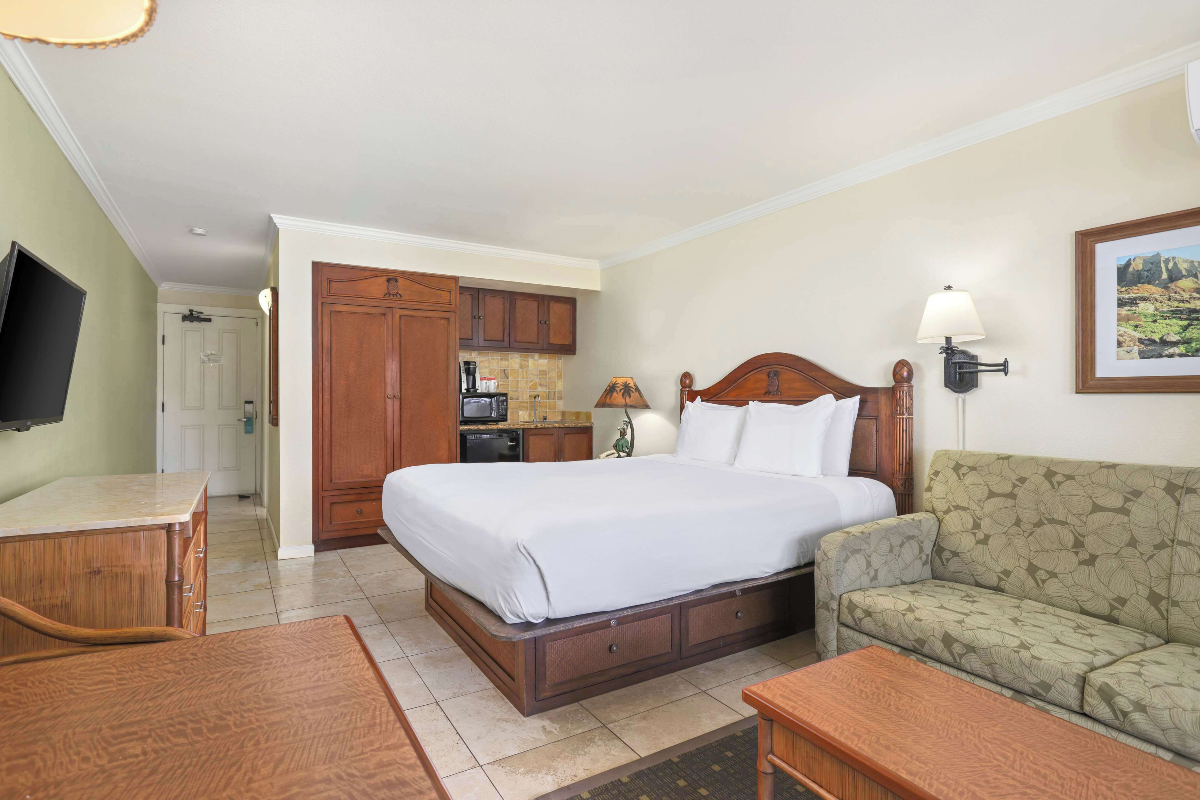 Quarto de hotel com cama, sofá, televisão, escrivaninha e cozinha compacta.