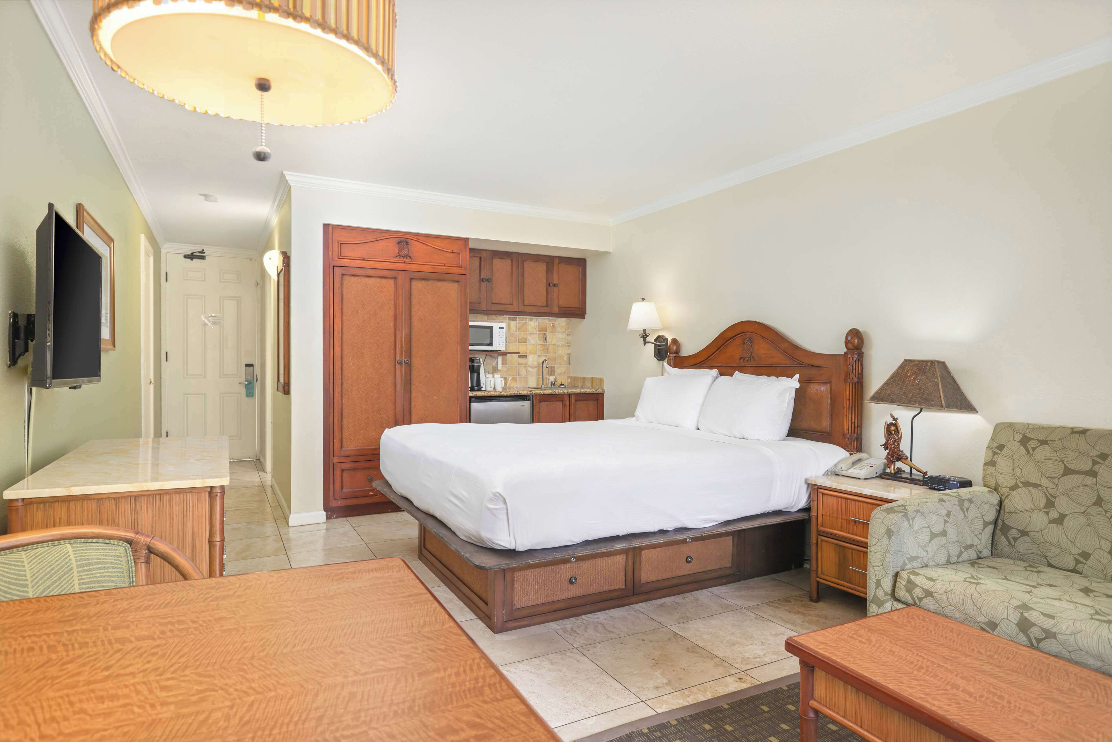 A hotel room with a bed, a desk, a chair, a sofa, a television, a lamp, and a kitchenette.